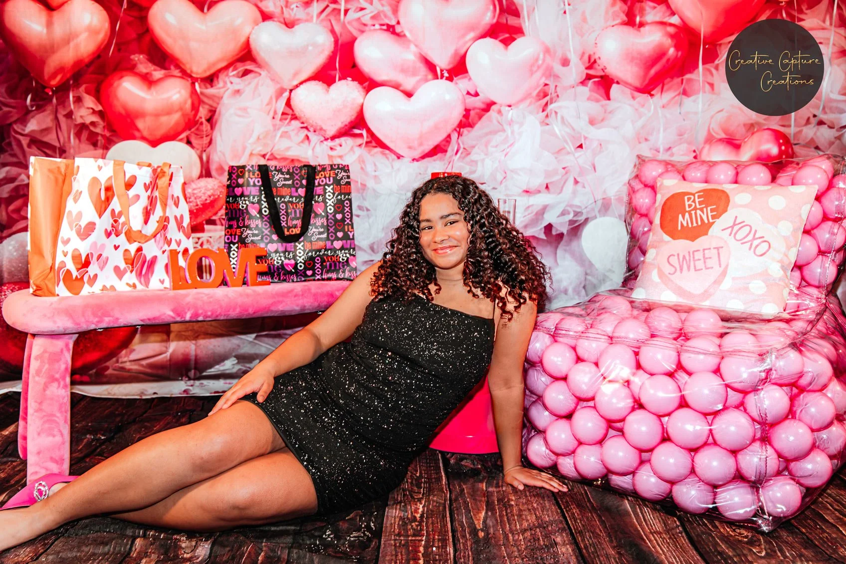 A woman with curly hair in a black dress sitting on the floor next to a pink chair filled with pink balls, surrounded by Valentine's Day decorations including balloons, gift bags, and a heart-themed backdrop.