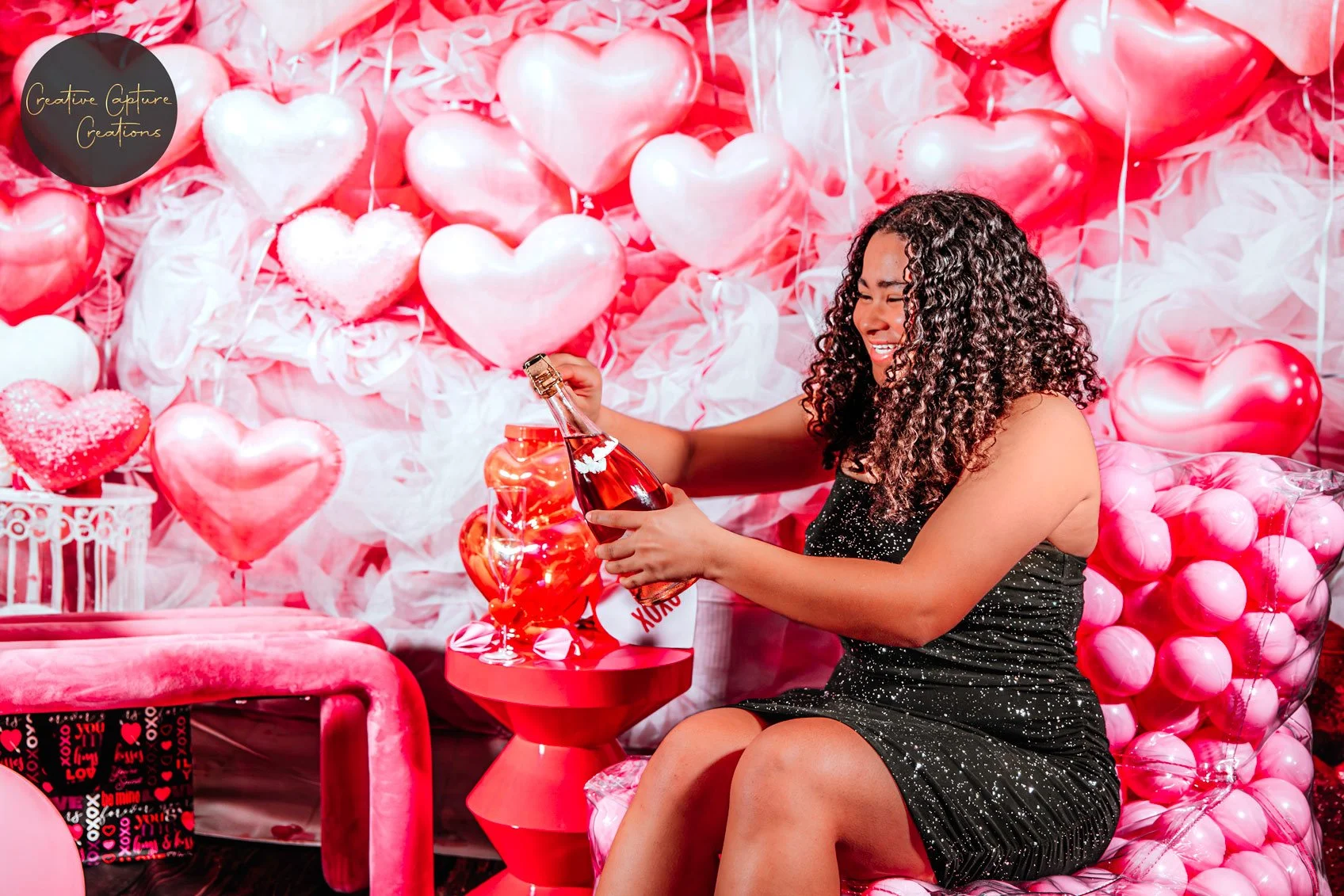 A woman with curly hair in a black dress opens a bottle of pink champagne in front of a heart-themed balloon backdrop.