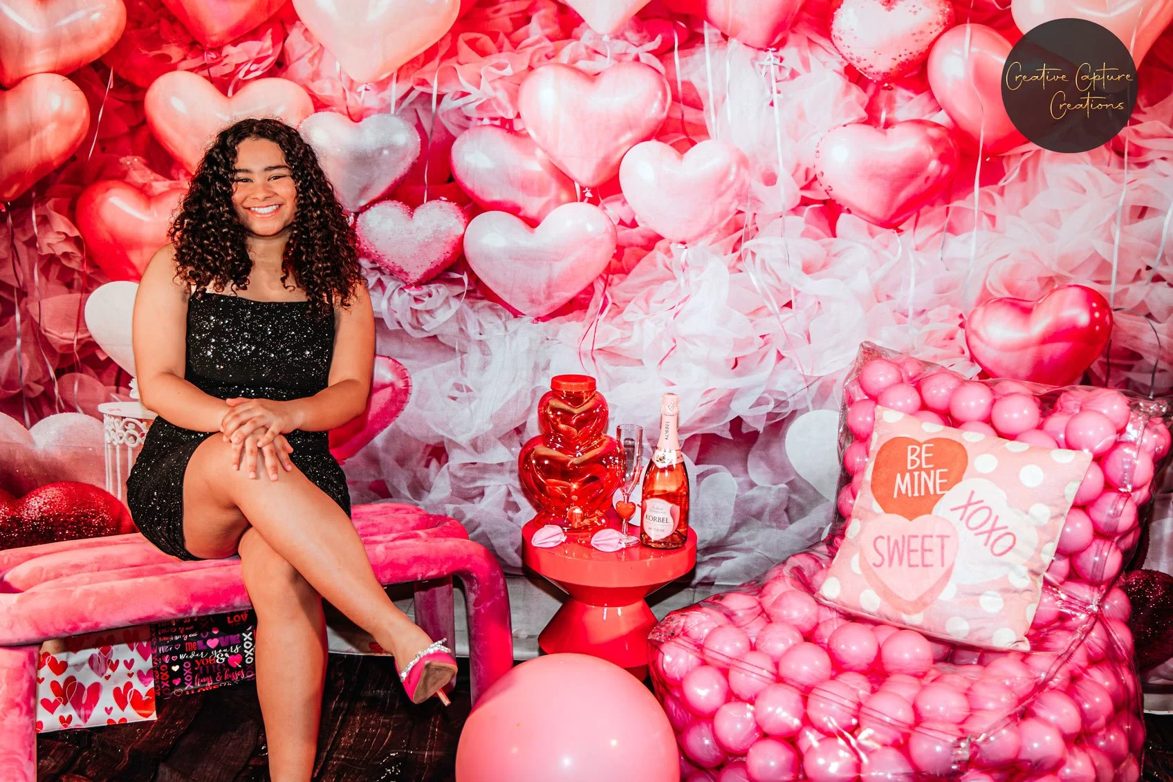 A woman sitting on a pink velvet bench in front of a Valentine-themed backdrop with numerous pink, red, and white heart-shaped balloons and pink tissue paper. She is smiling, wearing a black sparkly dress, with her legs crossed and hands resting on h