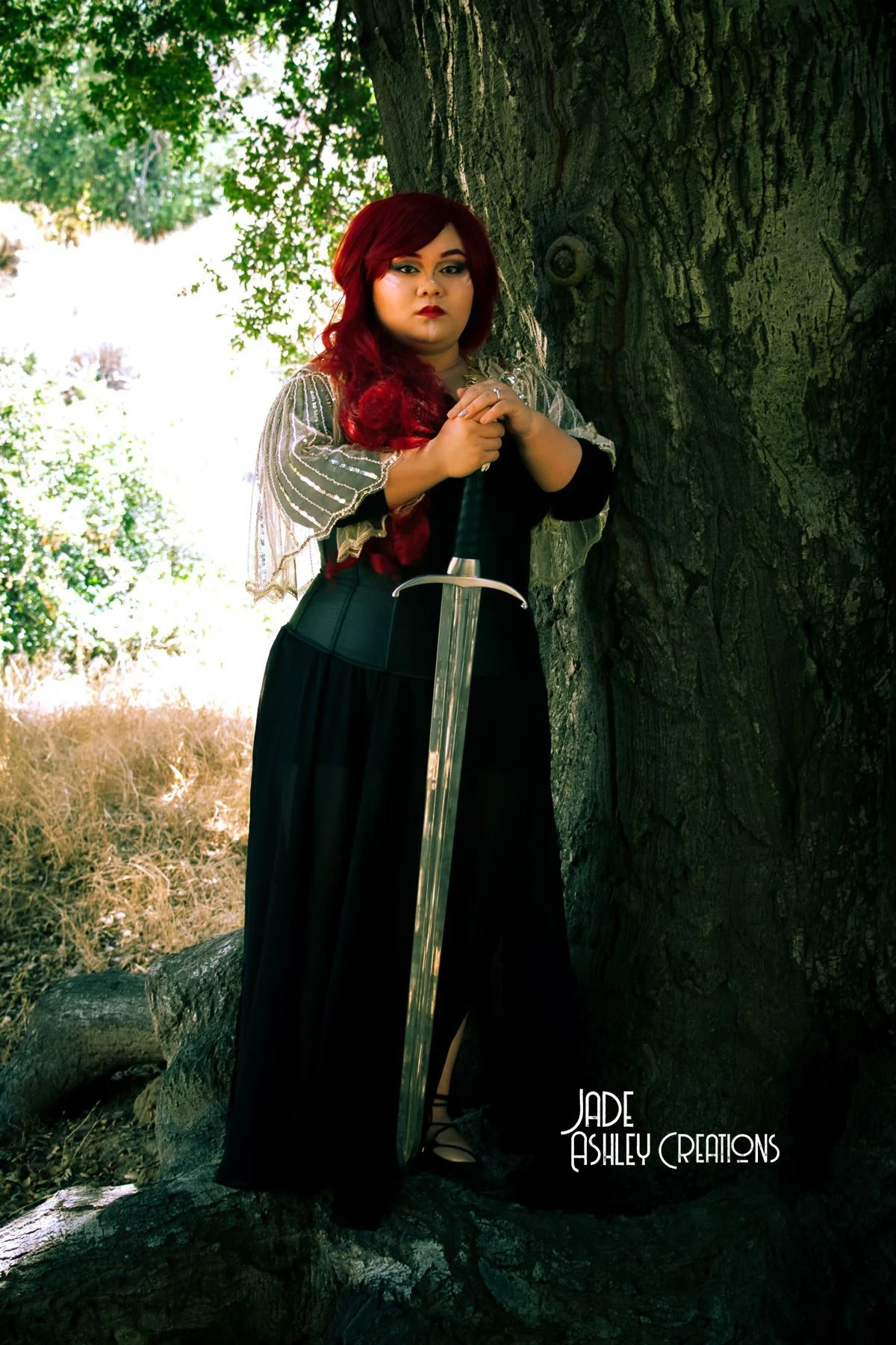 A woman with red hair and dark makeup leaning against a large tree trunk in a forested area, holding a sword upright in front of her. She wears a black dress and a sheer, silver-embellished cape.