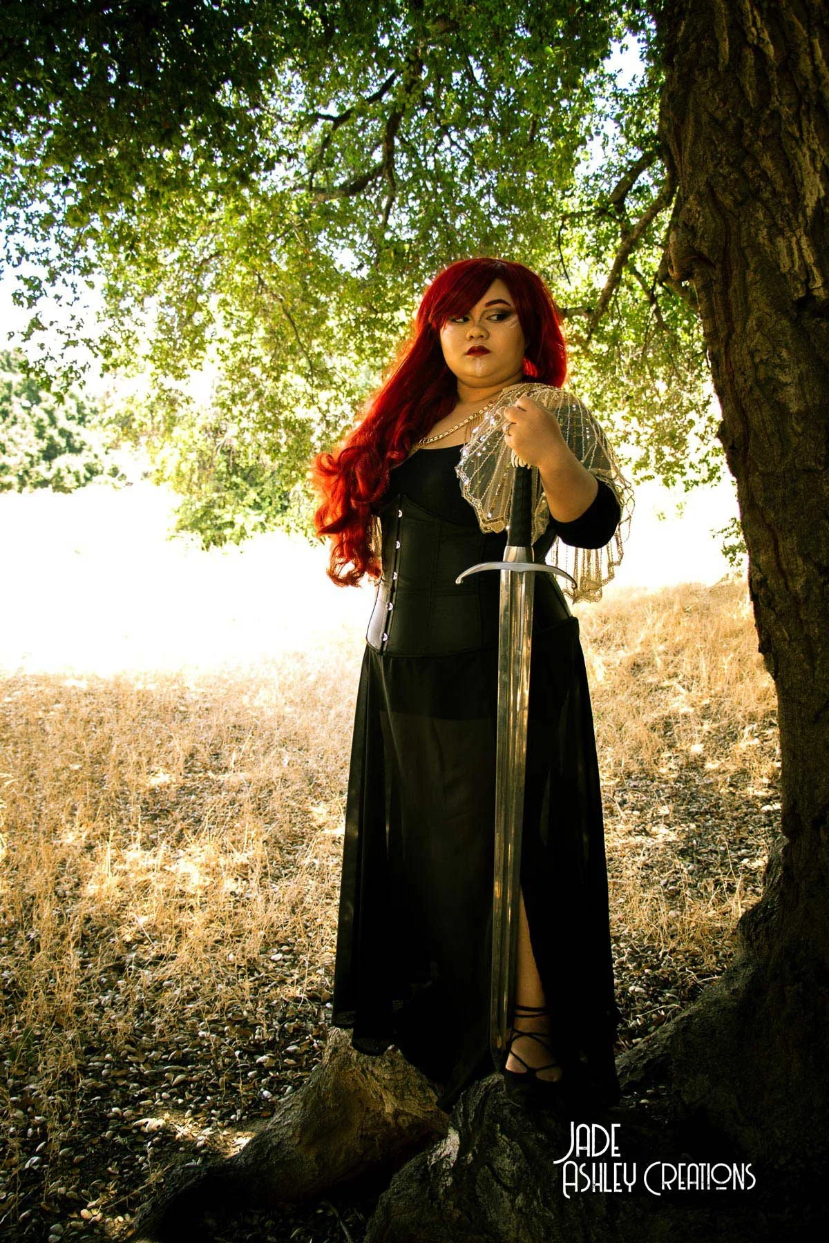 A woman with long red hair is standing next to a large tree, holding a sword upright in a forested area with partly green and partly dried grass. She is dressed in gothic style black clothing with lace and shiny accents.