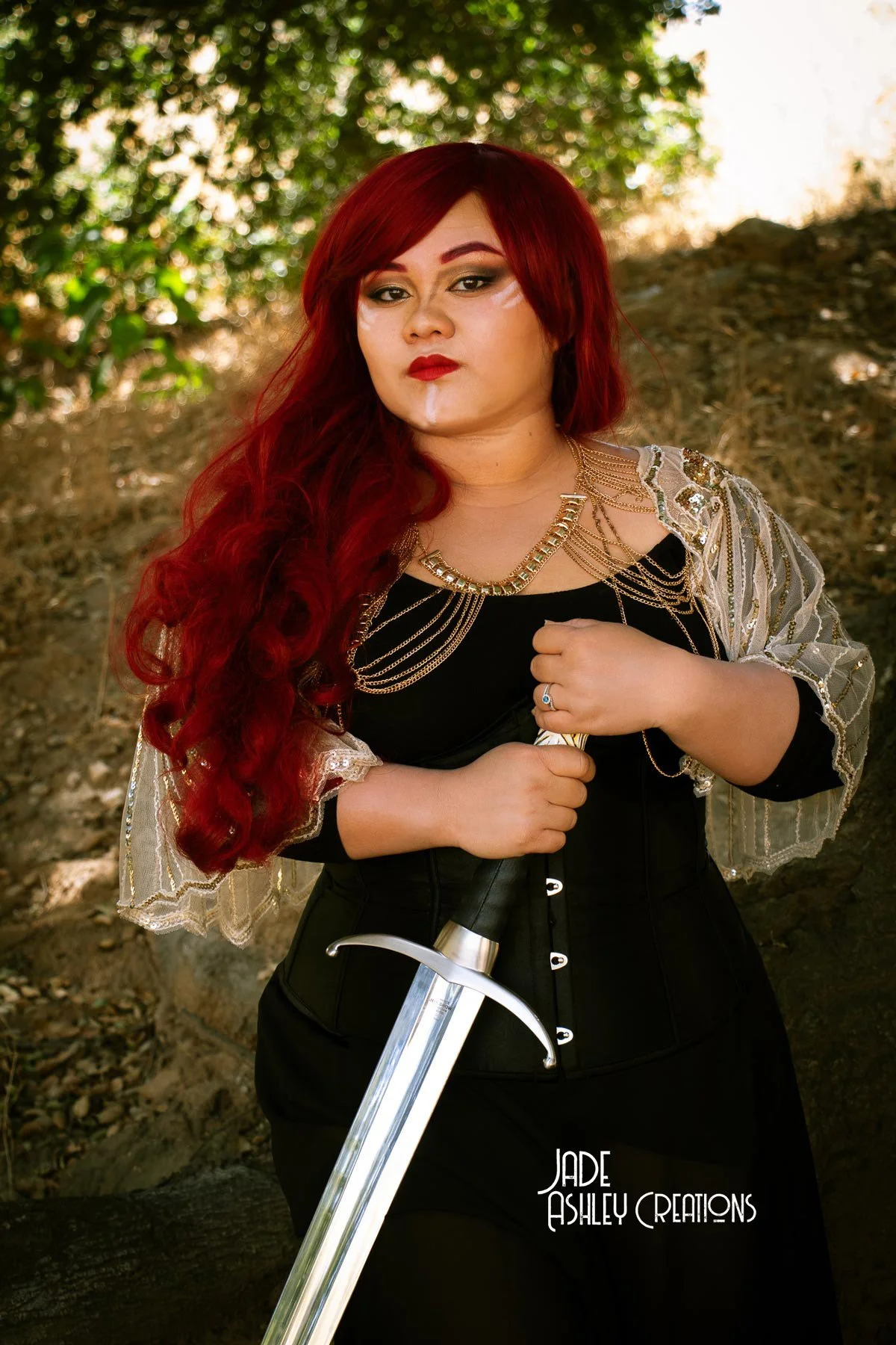 A woman with long, wavy red hair and elaborate makeup, wearing a black dress with gold jewelry and a silver, gold, and white lace overlay, holding a sword in an outdoor setting with trees in the background.