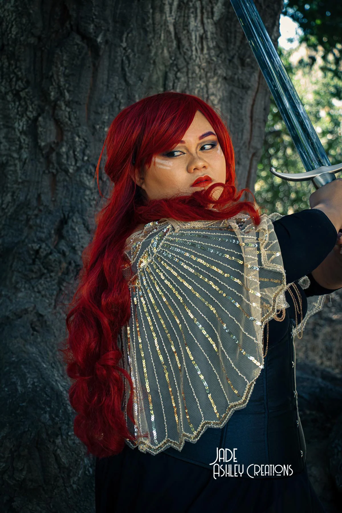 Woman with long red hair and fierce expression, wearing a detailed, shiny cape and holding a sword, standing outdoors in front of a large tree.