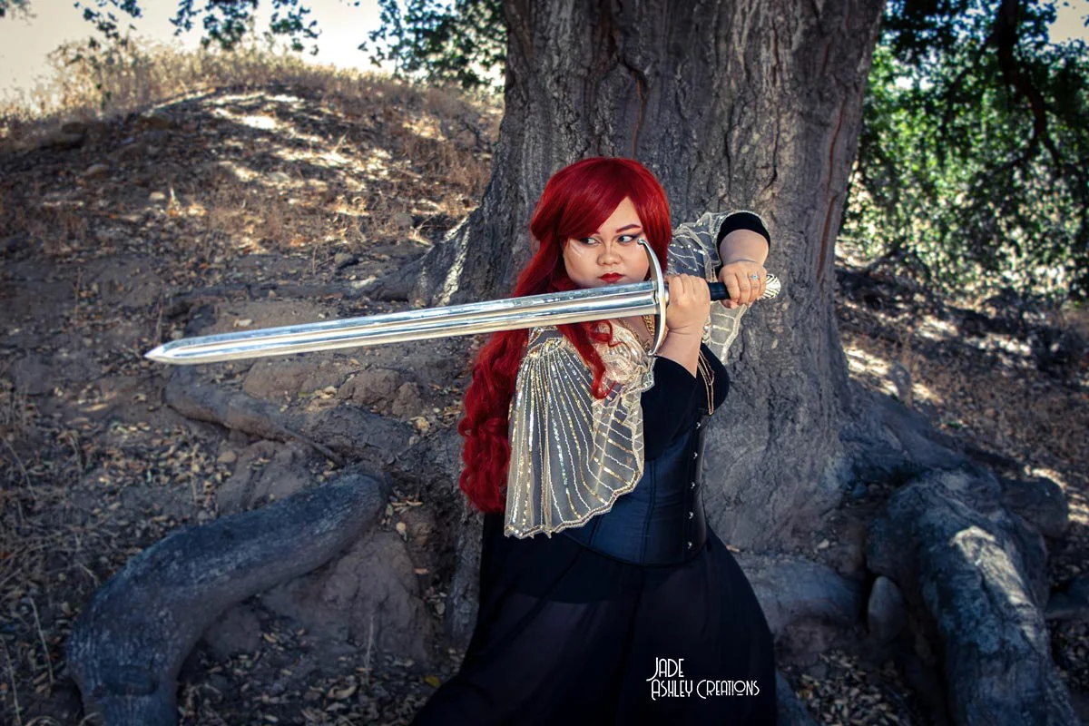 A woman with long red hair, dressed in a black outfit with a decorated sheer cape, holding a large sword in a combat stance outdoors near a tree with a rocky, dirt ground.
