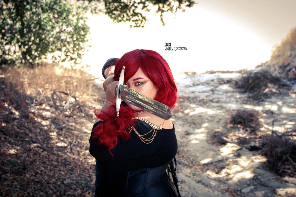 A woman with bright red hair, wearing black clothing with gold chains on her shoulder, holds a sword in front of her, standing on a dirt path in an outdoor setting with trees and sunlight.