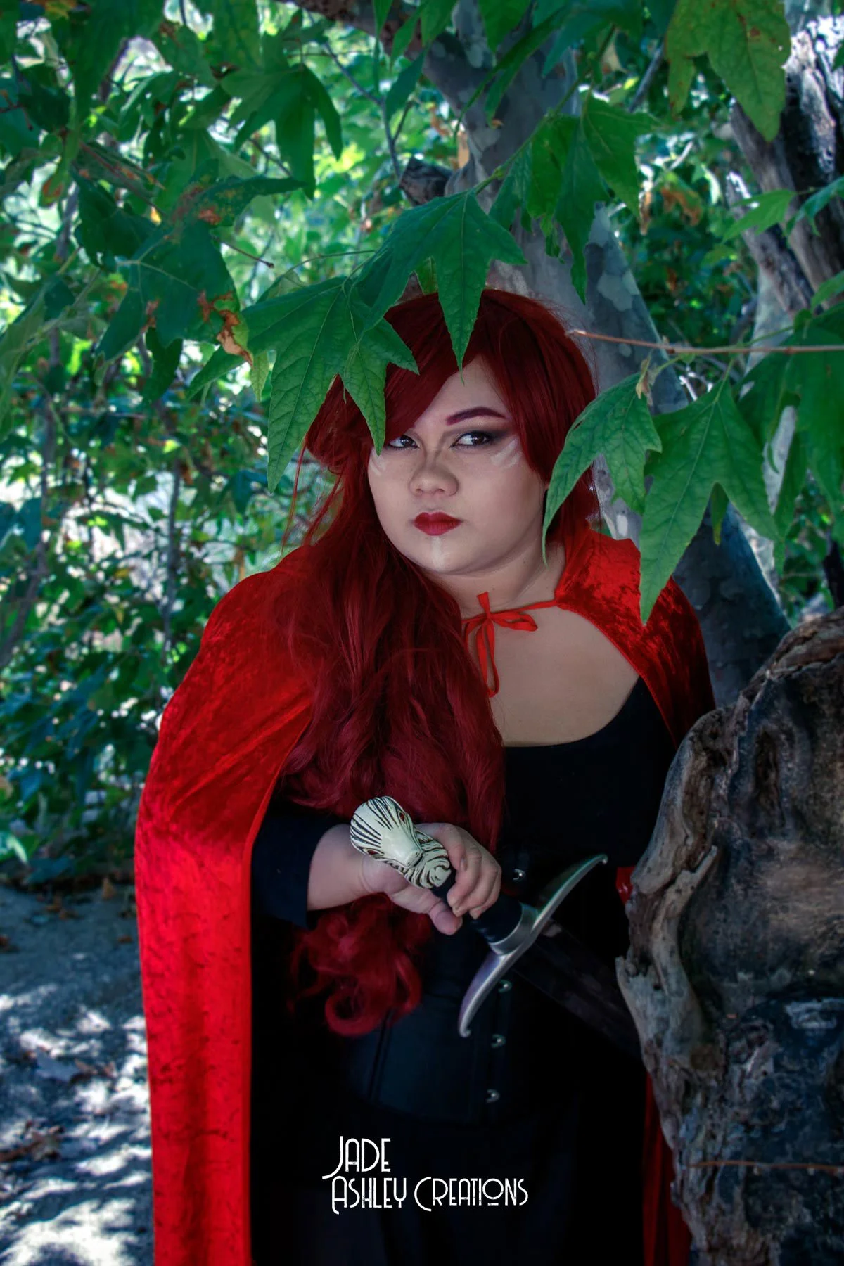 A woman with long red hair dressed as a princess or fairy tale character, wearing a red cape and holding a dagger, stands behind a tree with green leaves, in a forest setting.