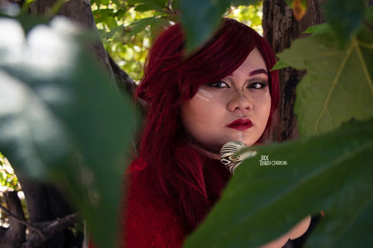 A woman with long red hair and makeup posing outdoors behind large green leaves with a tree trunk in the background.