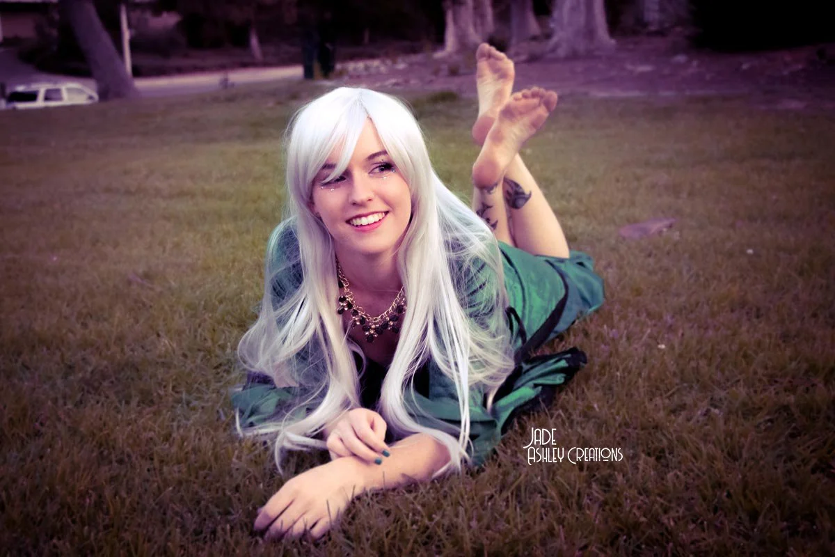 A woman with long platinum blonde hair lying on her stomach on a grassy field, smiling and looking to the side, with trees and a car in the background.