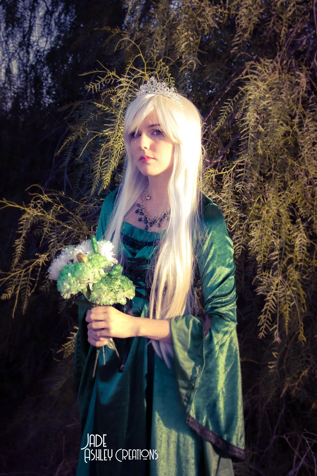 A woman dressed as a fantasy queen or fairy with long platinum blonde hair, wearing a tiara, a green velvet gown, and a necklace, holding a bouquet of white and green flowers, standing in front of a bushy background with leafy branches.