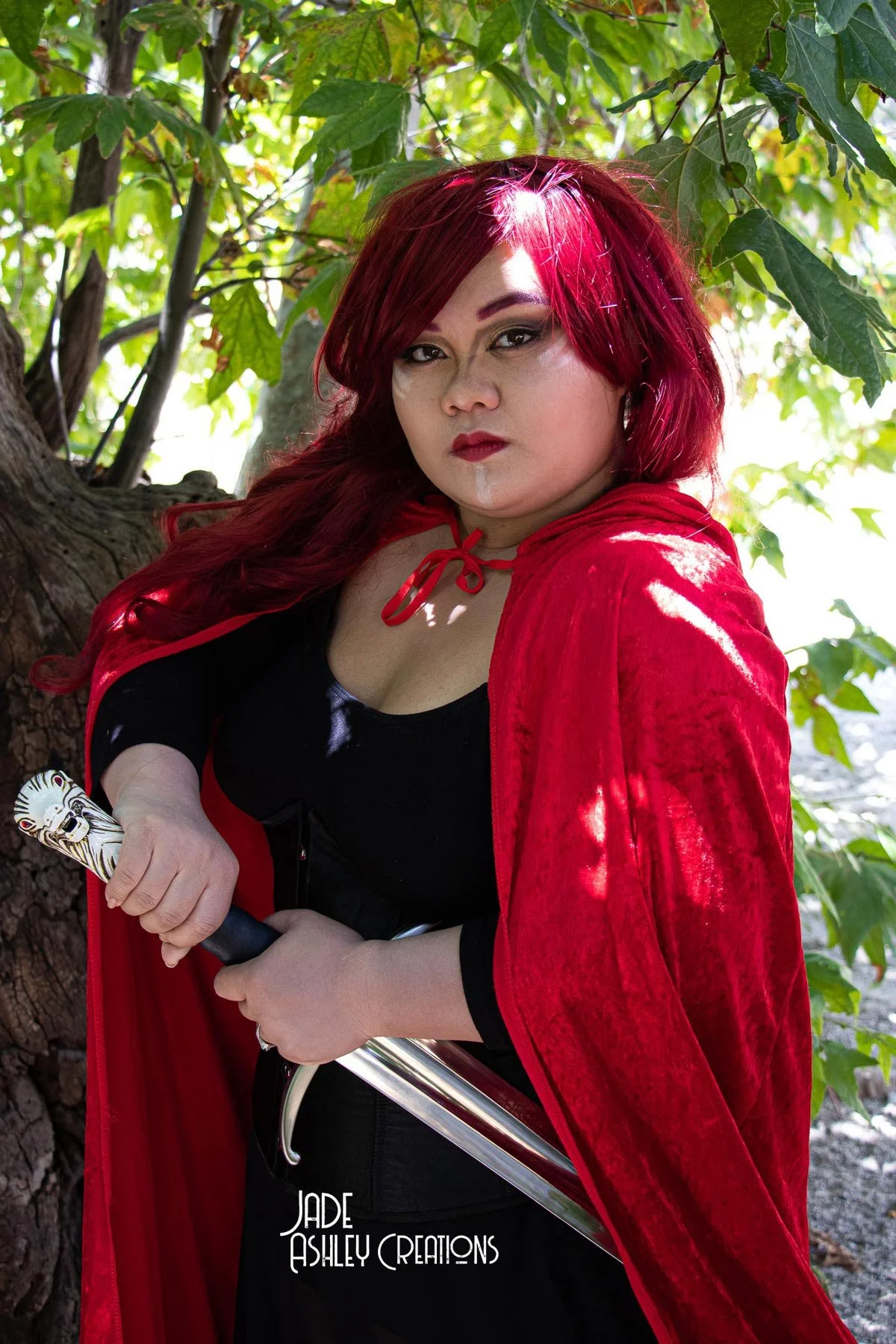 A woman dressed as a vampire princess with red hair and a red cloak, holding a sword, standing outdoors near a tree with green leaves.
