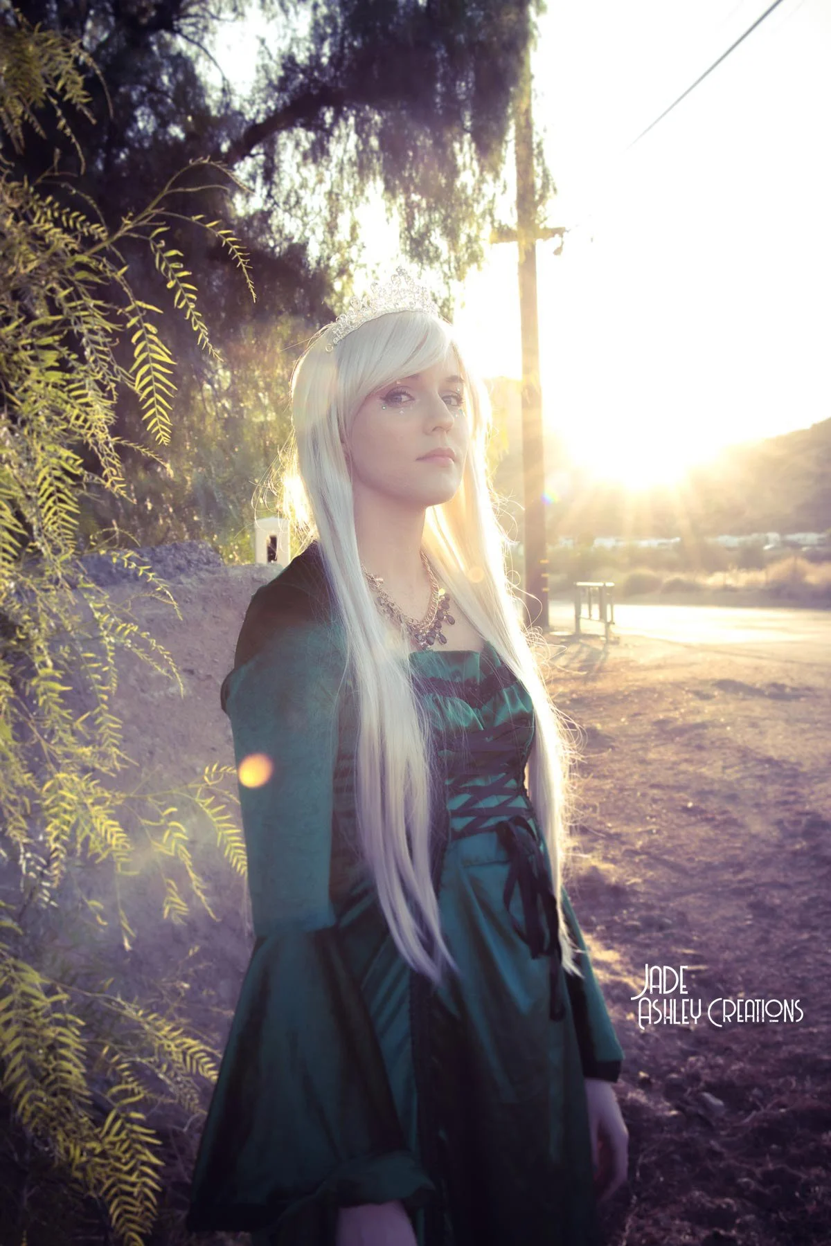A woman with long platinum blonde hair, wearing a tiara and a dark green corset-style dress, standing outdoors in a wooded area during sunset with trees and a dirt road in the background.