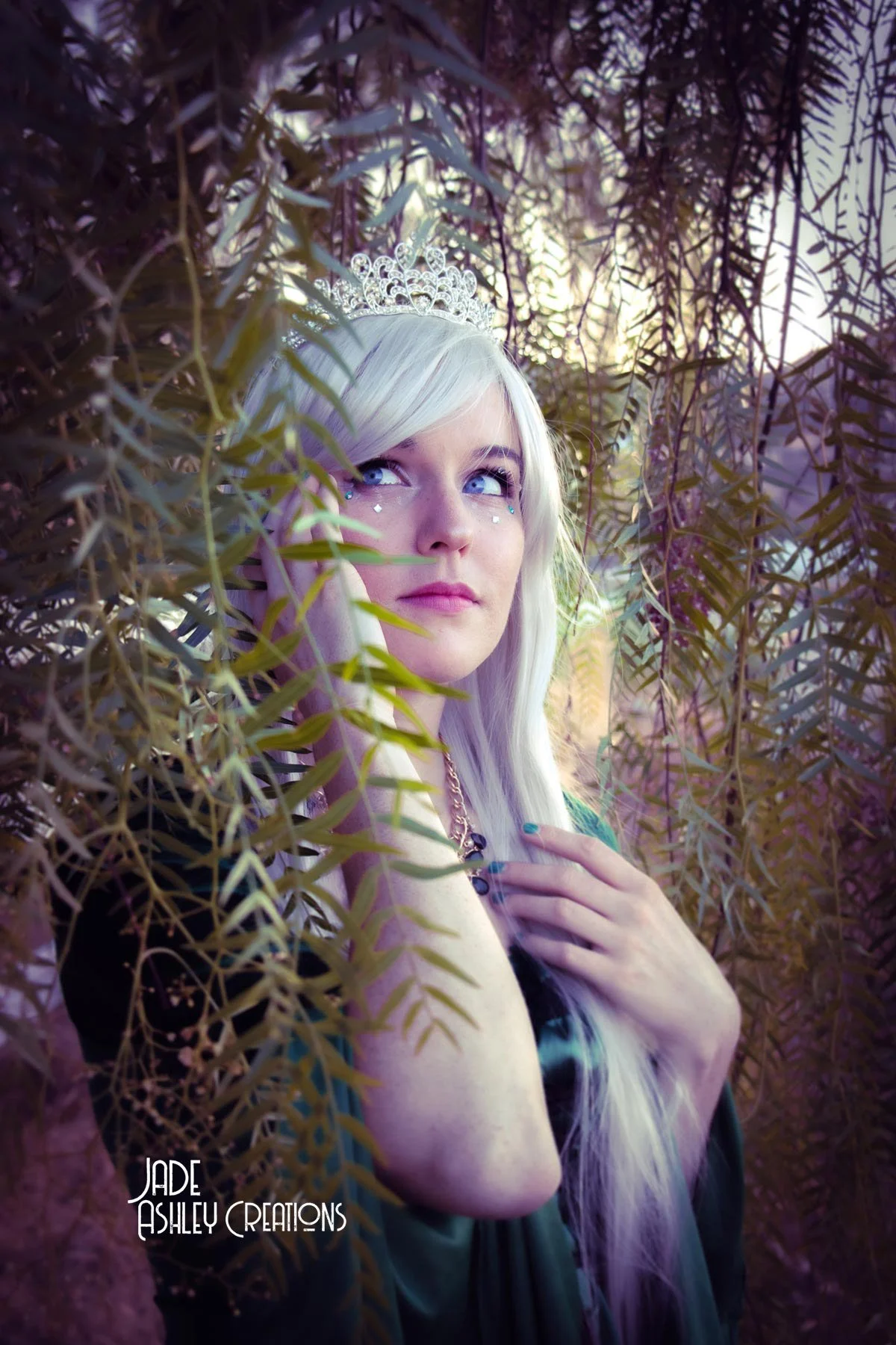 A woman with platinum blonde hair and a small tiara, looking thoughtfully through purple-tinted leaves, wearing a dark dress and a necklace, with the sunlight filtering through the foliage in the background.