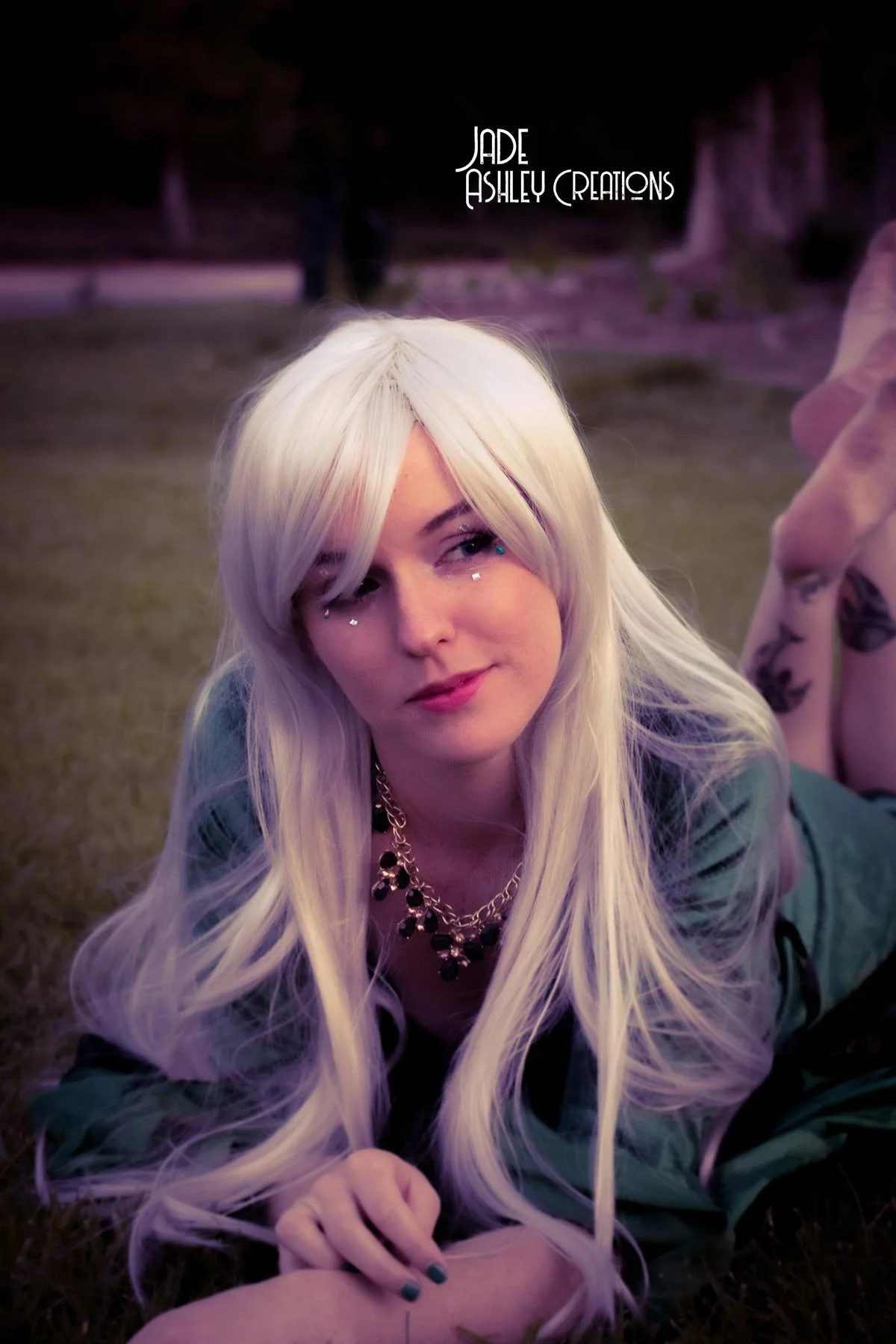A woman with long platinum blonde hair lying on her stomach on the grass, wearing a green jacket, black top, and a black and gold necklace, with small rhinestone decorations near her eyes, with a blurred outdoor background and text reading 'Jade Ashl