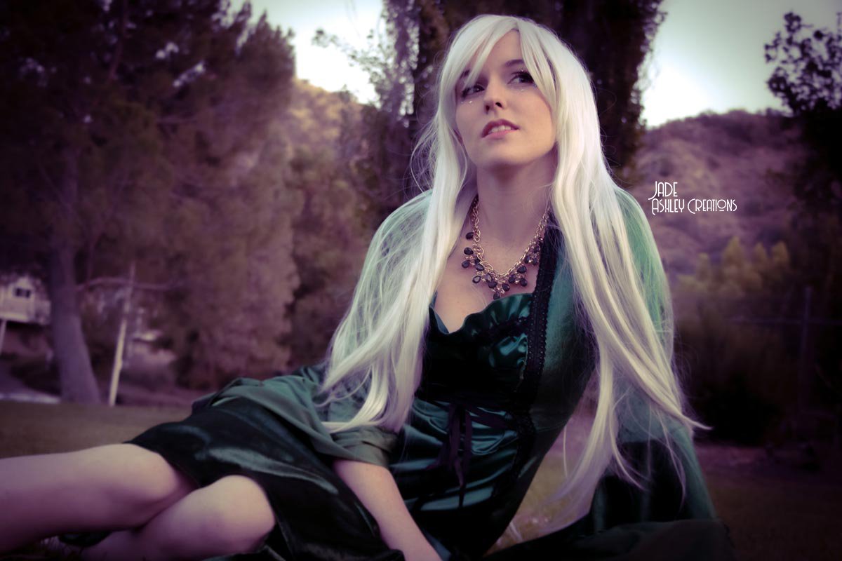 A woman with long blonde hair wearing a dark dress and statement necklace outdoors with trees and hills in the background.