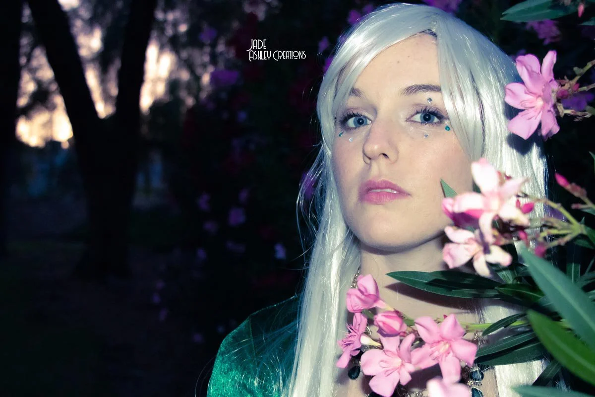 A woman with platinum blonde hair and blue eyes surrounded by pink flowers, in a dark natural setting at dusk.