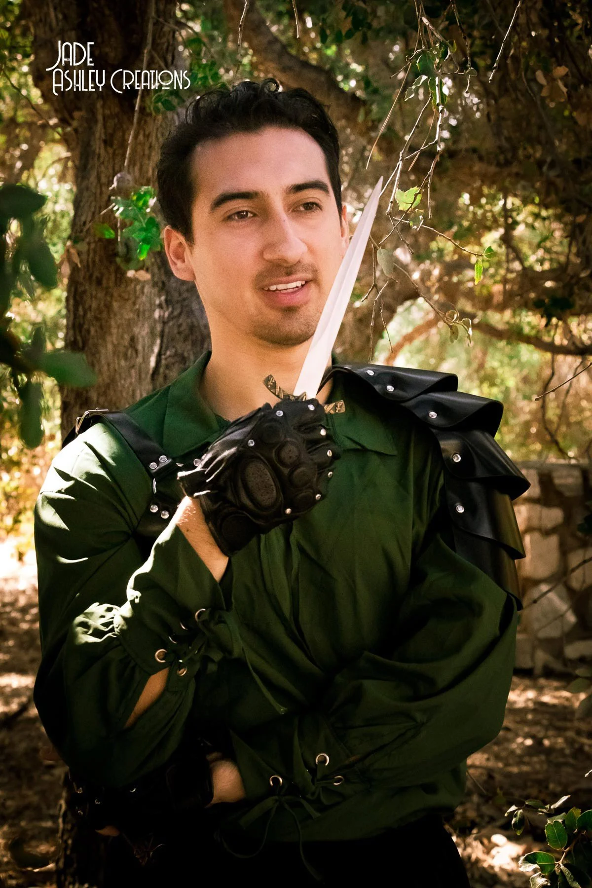A man in medieval fantasy armor, holding a dagger, standing outdoors in a wooded area.