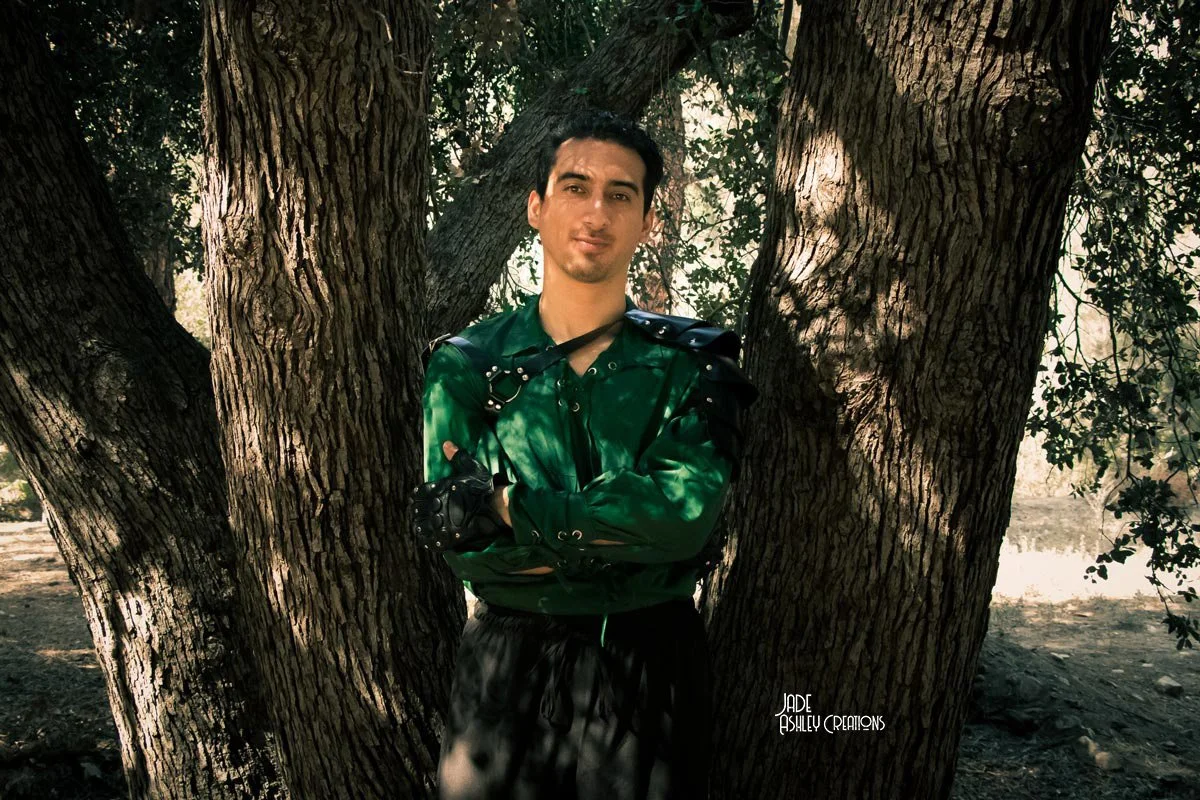 A young man with dark hair and a goatee standing between two large tree trunks outdoors, wearing a green leather jacket and black gloves, crossing his arms and looking at the camera with a slight smile.