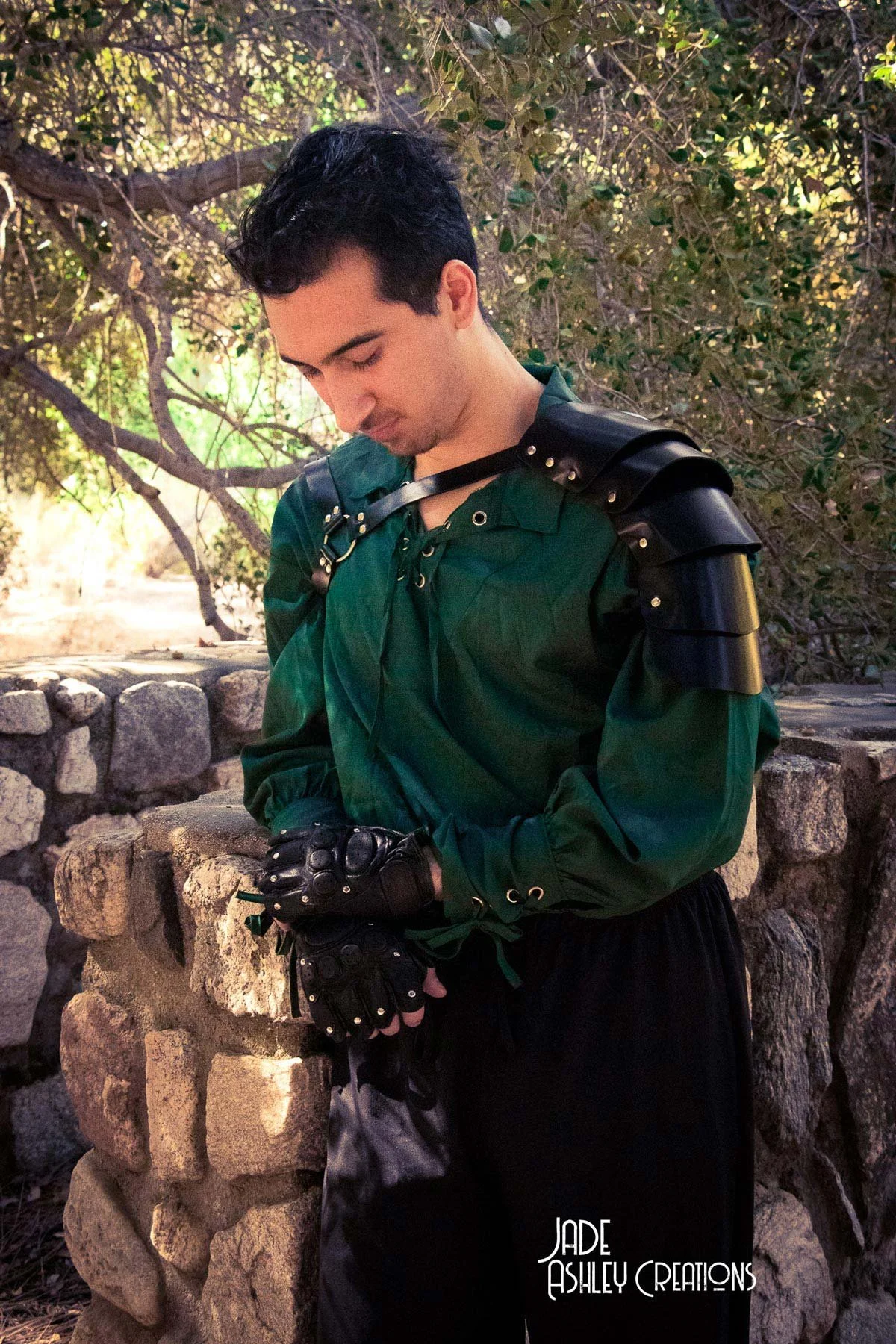 A young man dressed in a green shirt and black gloves with spiked armor on his shoulders, standing outdoors next to a stone wall with trees in the background, looking down.