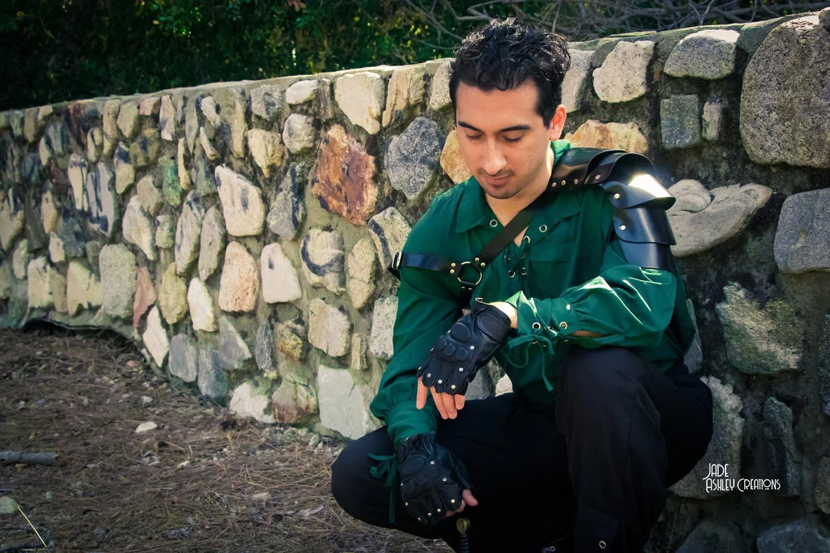 A young man dressed in a green shirt with black armor pads on his shoulder and a sleeve guard, crouches next to a stone wall outdoors, looking at his watch.