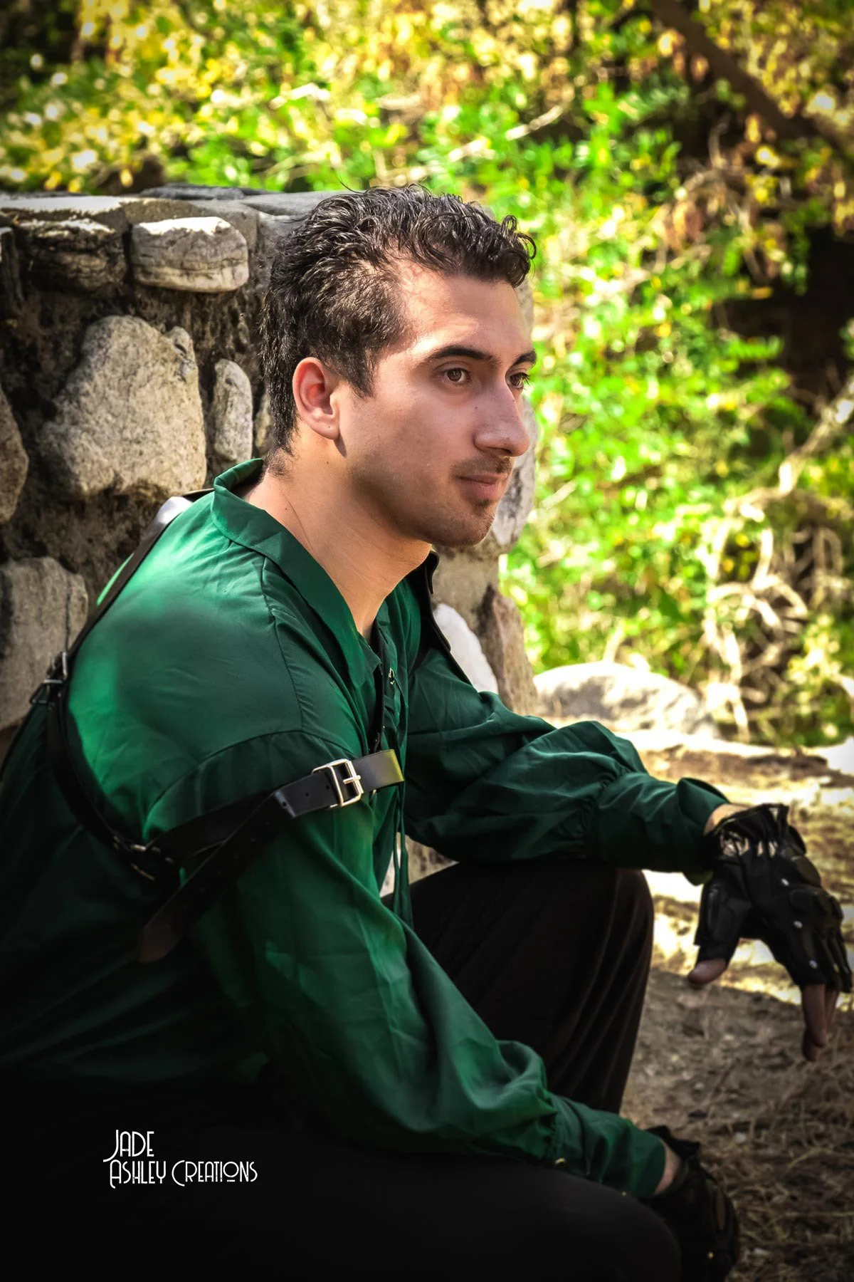 A young man with dark hair and light skin, sitting outdoors near a stone wall, wearing a green jacket and black gloves, with a backpack strap over his shoulder, looking thoughtfully into the distance, surrounded by greenery.