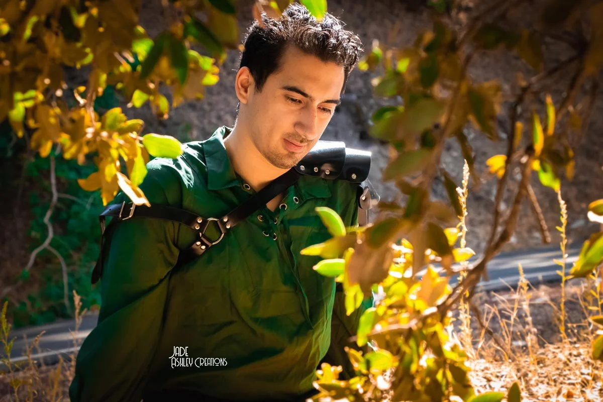 A young man with dark hair, wearing a green shirt and a black shoulder strap, looking down at bushes with green and brown leaves in an outdoor setting with sunlight.