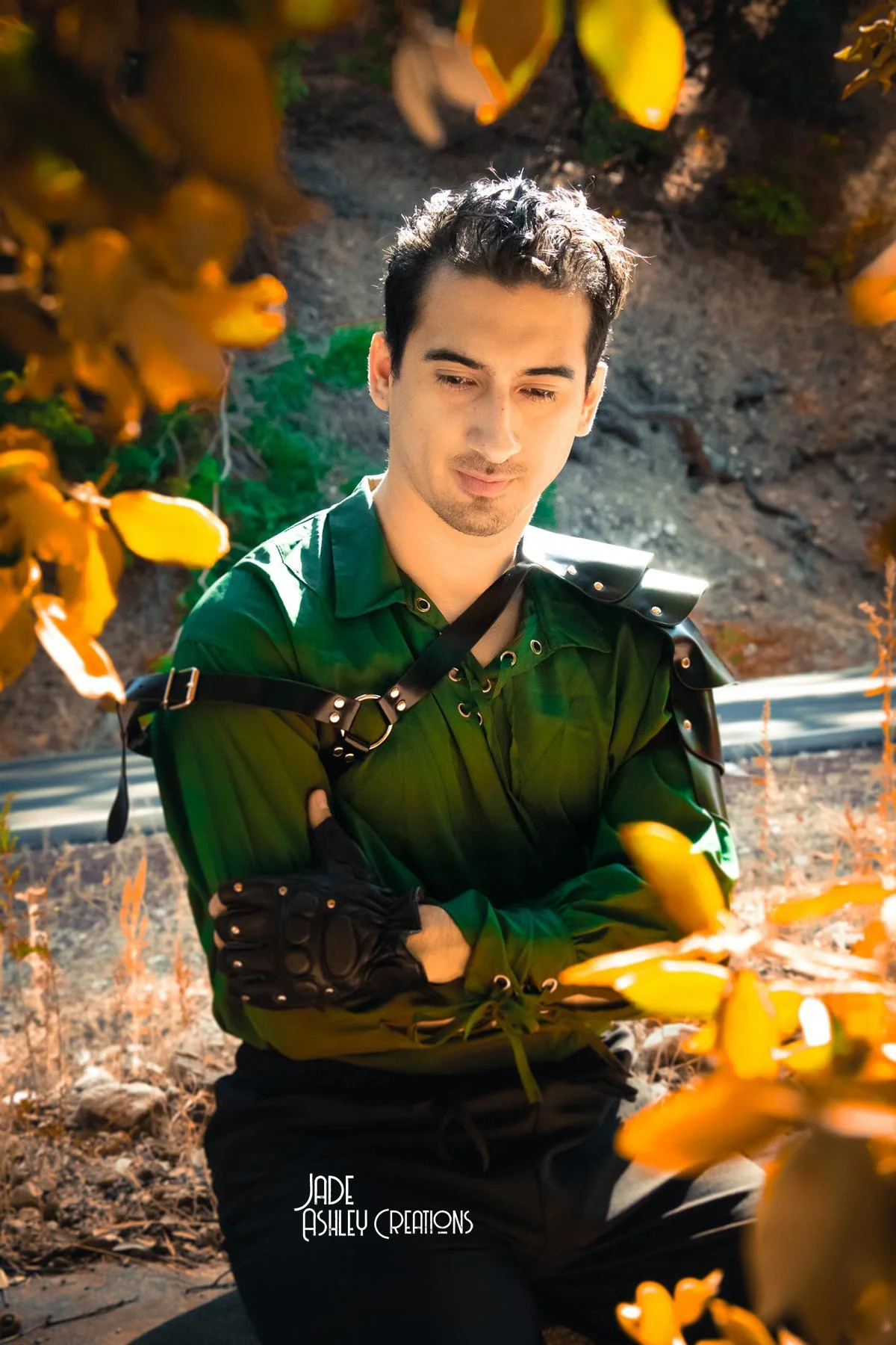 Young man with dark hair, wearing a green shirt and black gloves, stands outdoors among yellow-orange foliage with a mountainous background, crossing his arms and looking downward. The image has a watermark at the bottom reading "Jade Ashley Creation
