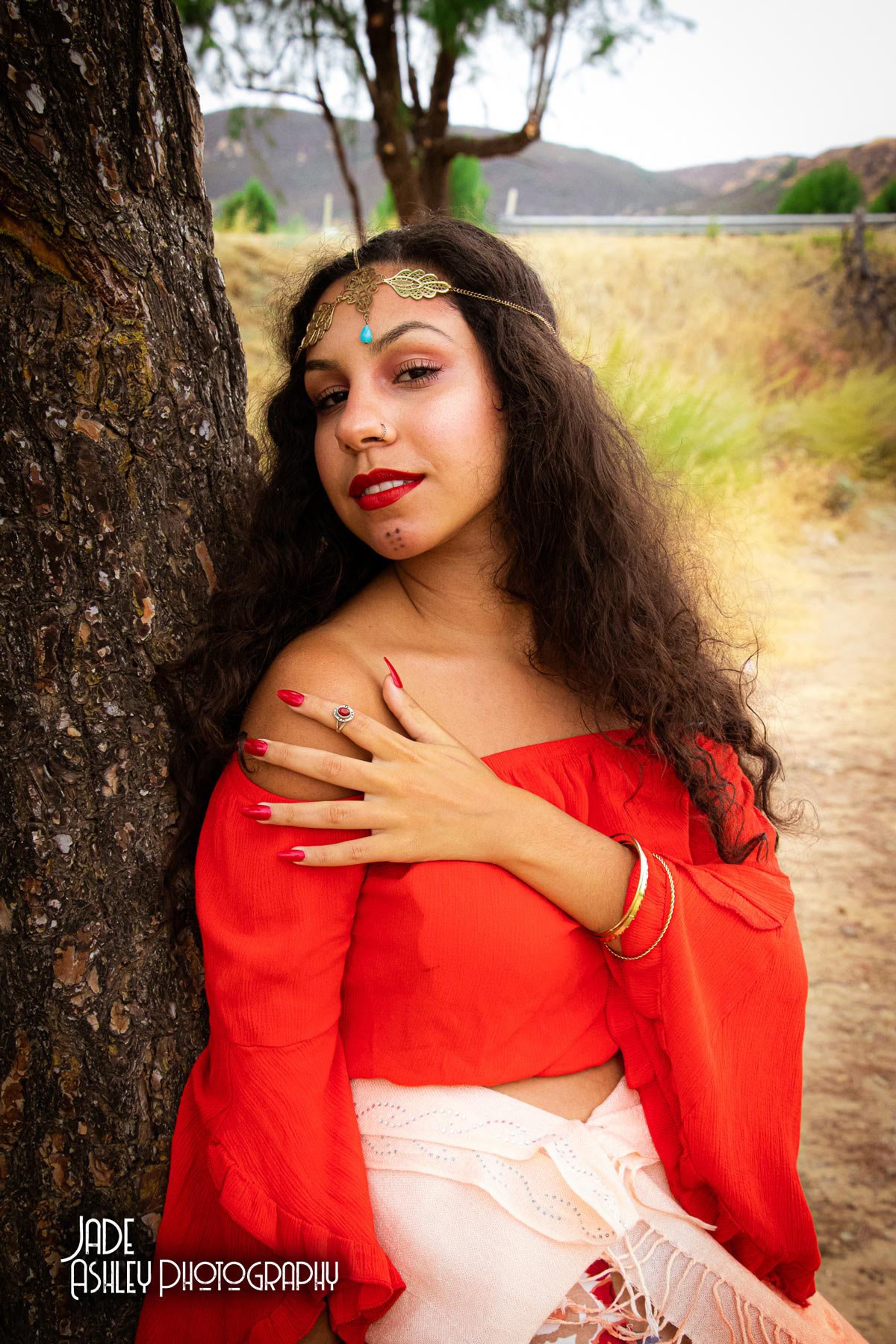 A woman with curly dark hair and red lipstick leaning against a tree outdoors. She is wearing a red off-shoulder top, torn white pants, and gold jewelry, including a headpiece, rings, and bracelets. The background shows a dirt path, trees, and hills.