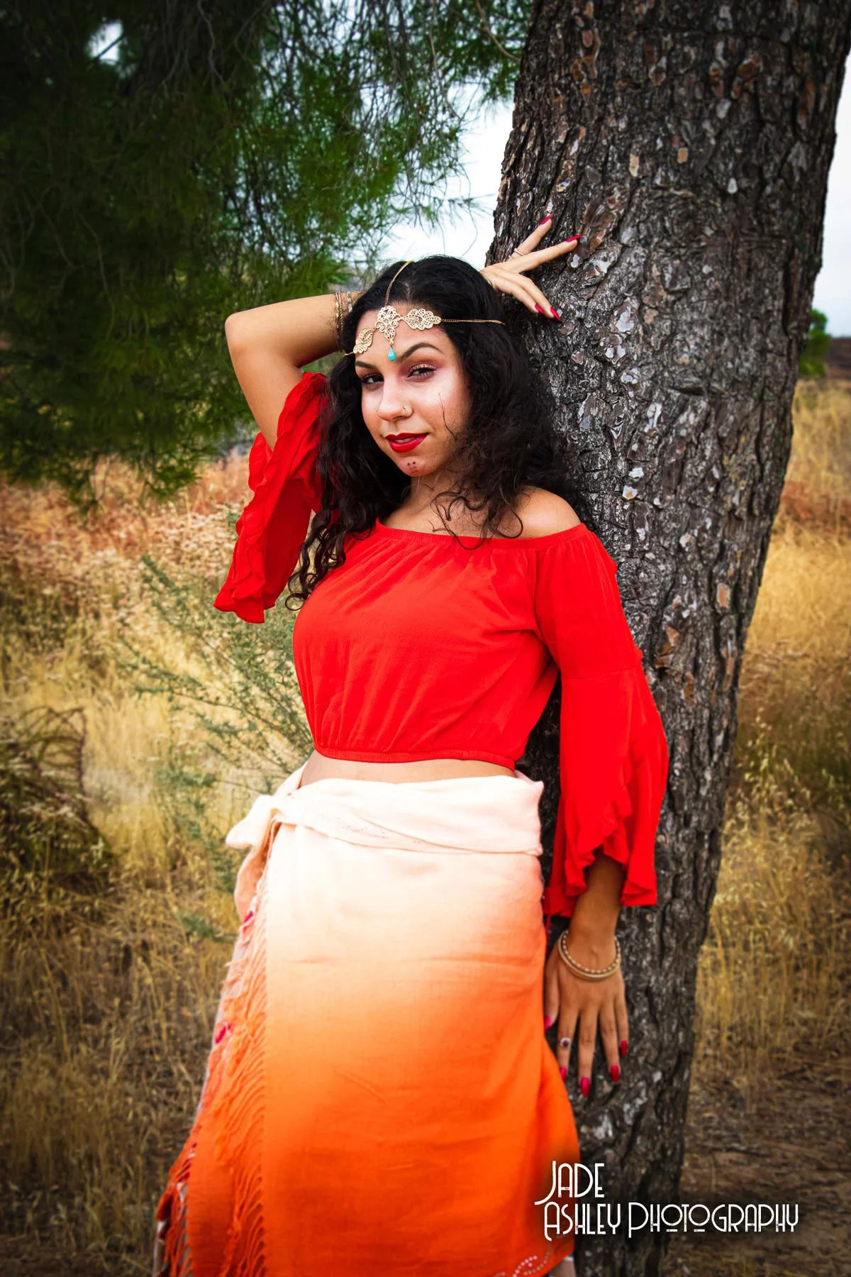 A woman dressed in a vibrant red off-shoulder top and an orange skirt, standing outdoors and leaning against a large tree, with a background of dry grass and green foliage.