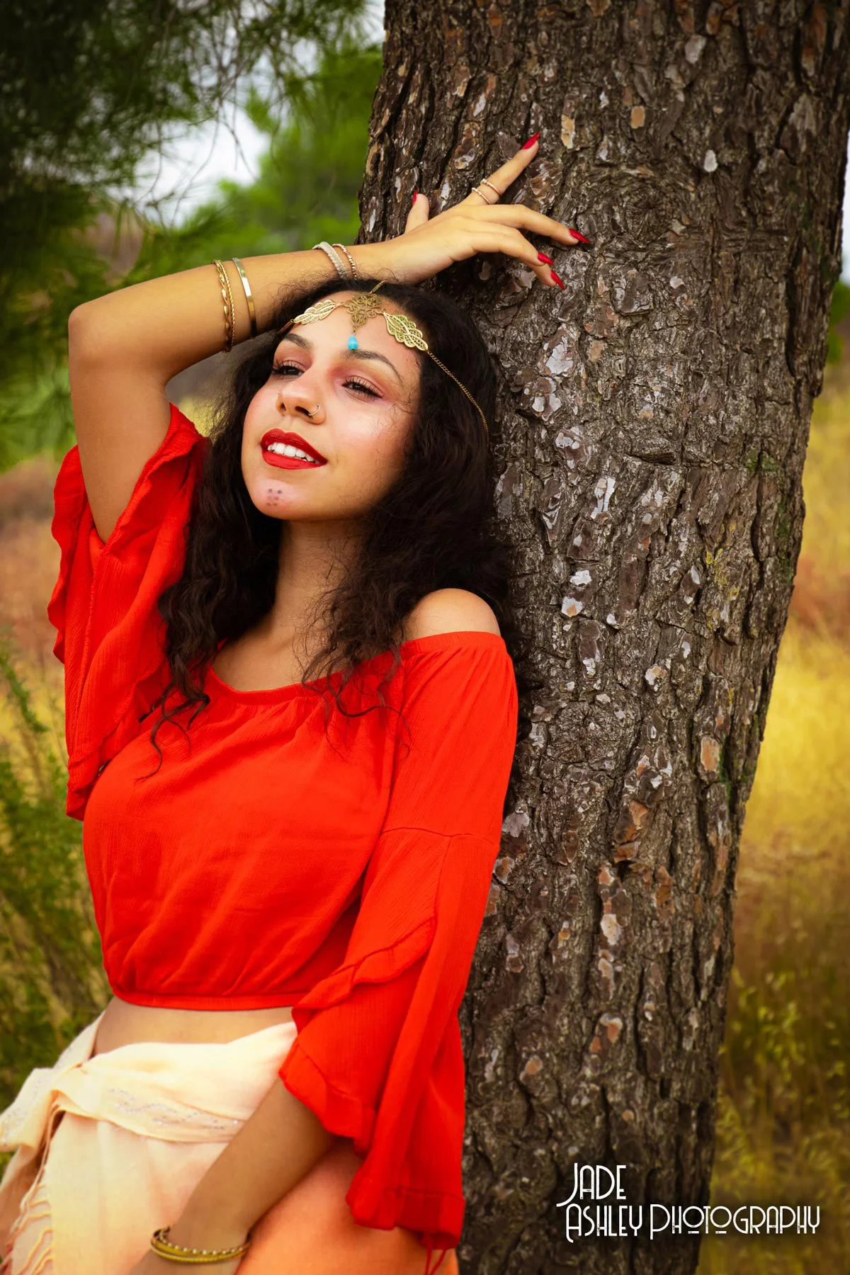 A woman with long black curly hair and red lipstick leaning against a tree in an outdoor setting. She is wearing a red off-shoulder top, beige skirt, and jewelry, including a head chain, bracelets, and rings. She has one arm raised, touching the tree