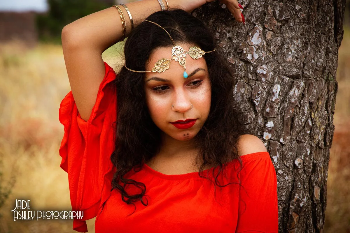 A woman with dark curly hair wearing a red off-the-shoulder top, bold red lipstick, a nasal piercing, and a decorative gold headpiece with a blue teardrop, leaning against a tree in a natural outdoor setting.