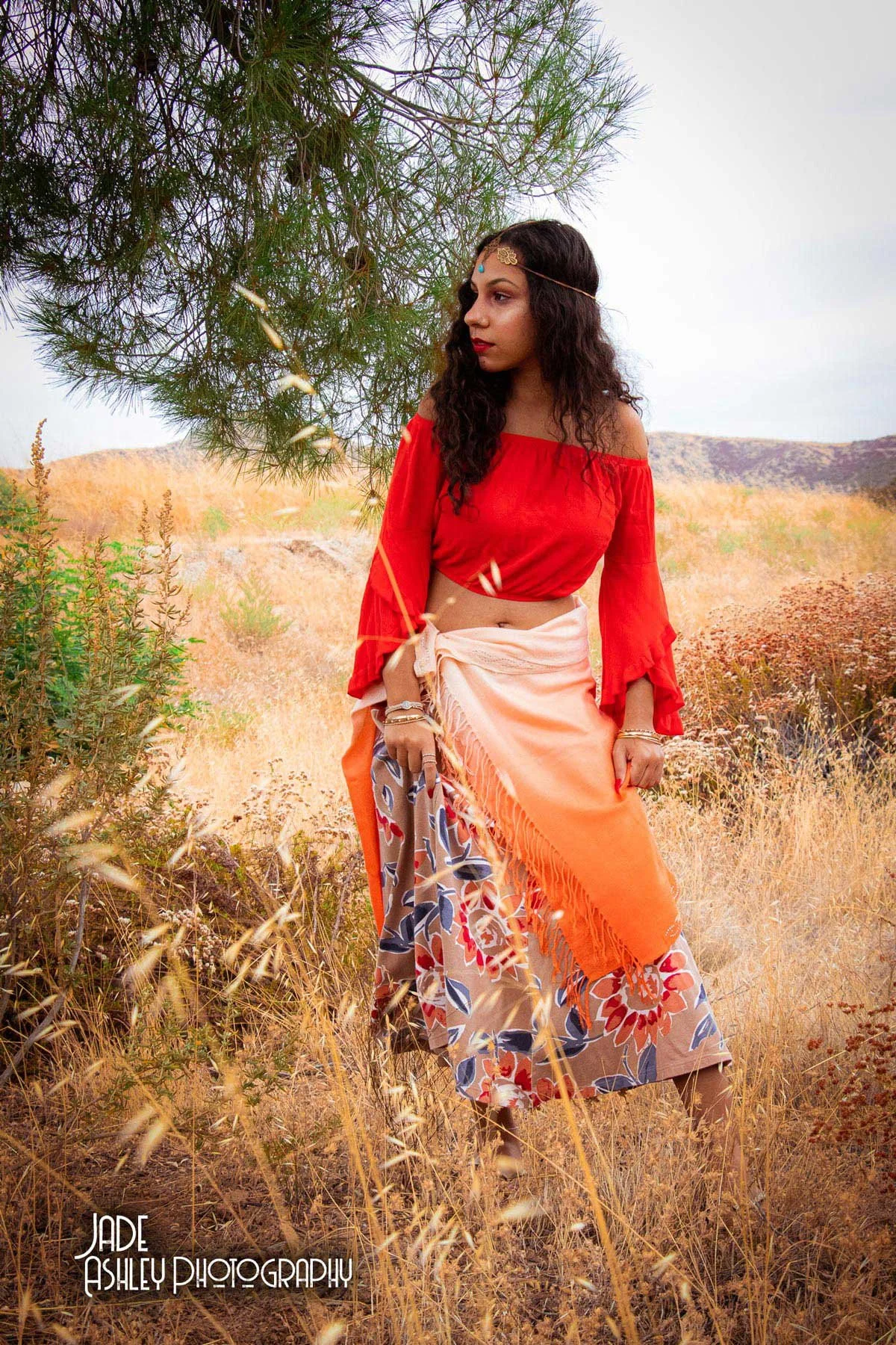A woman dressed in bohemian style clothing, wearing a red off-the-shoulder top, a floral skirt, and peach-colored shawl, standing in a dry grassy field with mountains in the background, under a large green tree.
