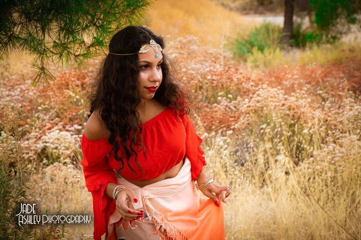 A woman with long, curly dark hair wearing a silver headpiece and red lipstick, dressed in a bright red off-shoulder top and a peach-colored skirt, standing outdoors among autumn foliage.