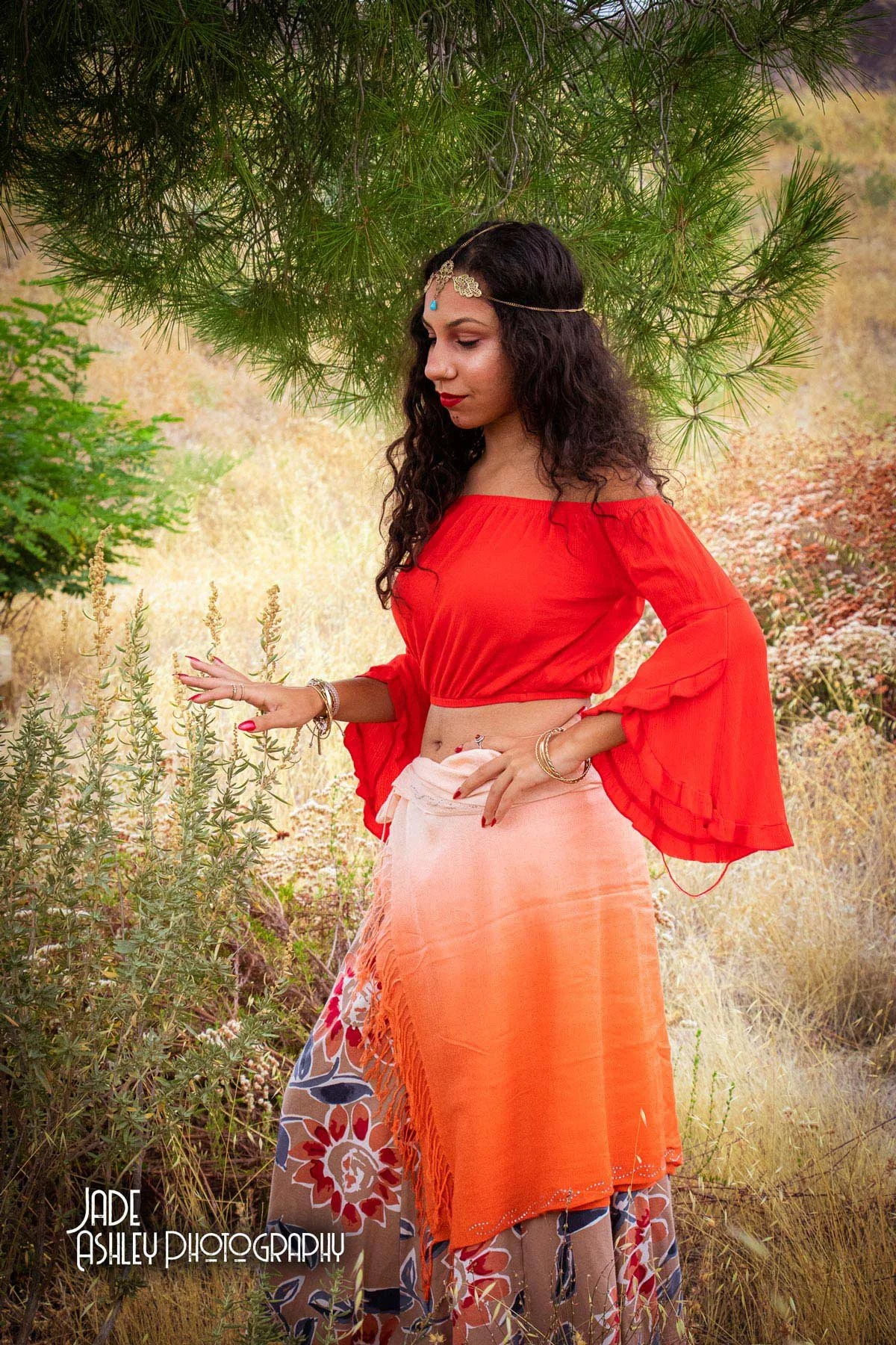 A woman with curly dark hair wearing a red off-shoulder top, an orange shawl, and a patterned skirt stands in a natural outdoor setting with foliage and grasses, softly touching plants under a tree.