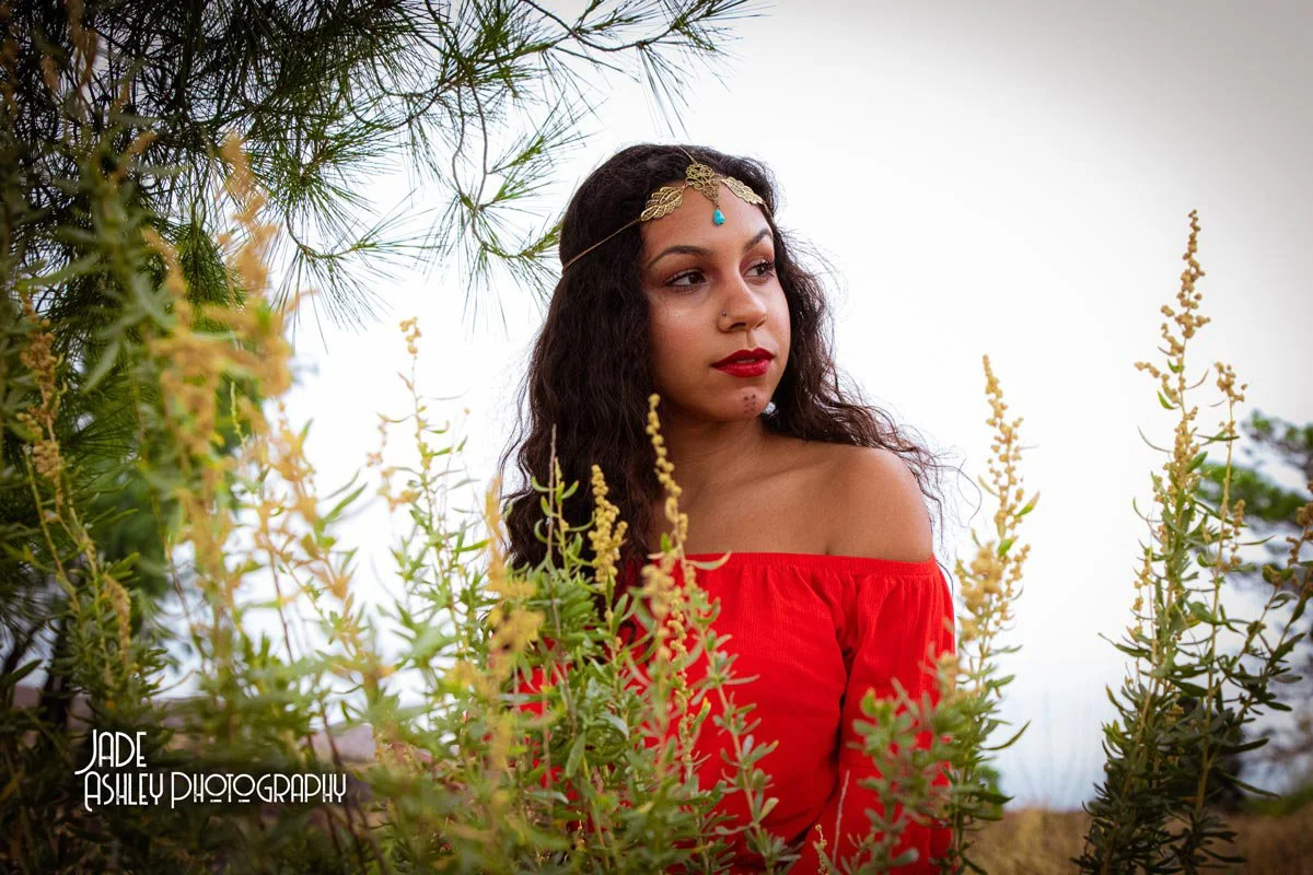 A woman with wavy dark hair, wearing a red off-shoulder top and jewelry with a blue gemstone, stands among yellow-green plants and trees during daylight.