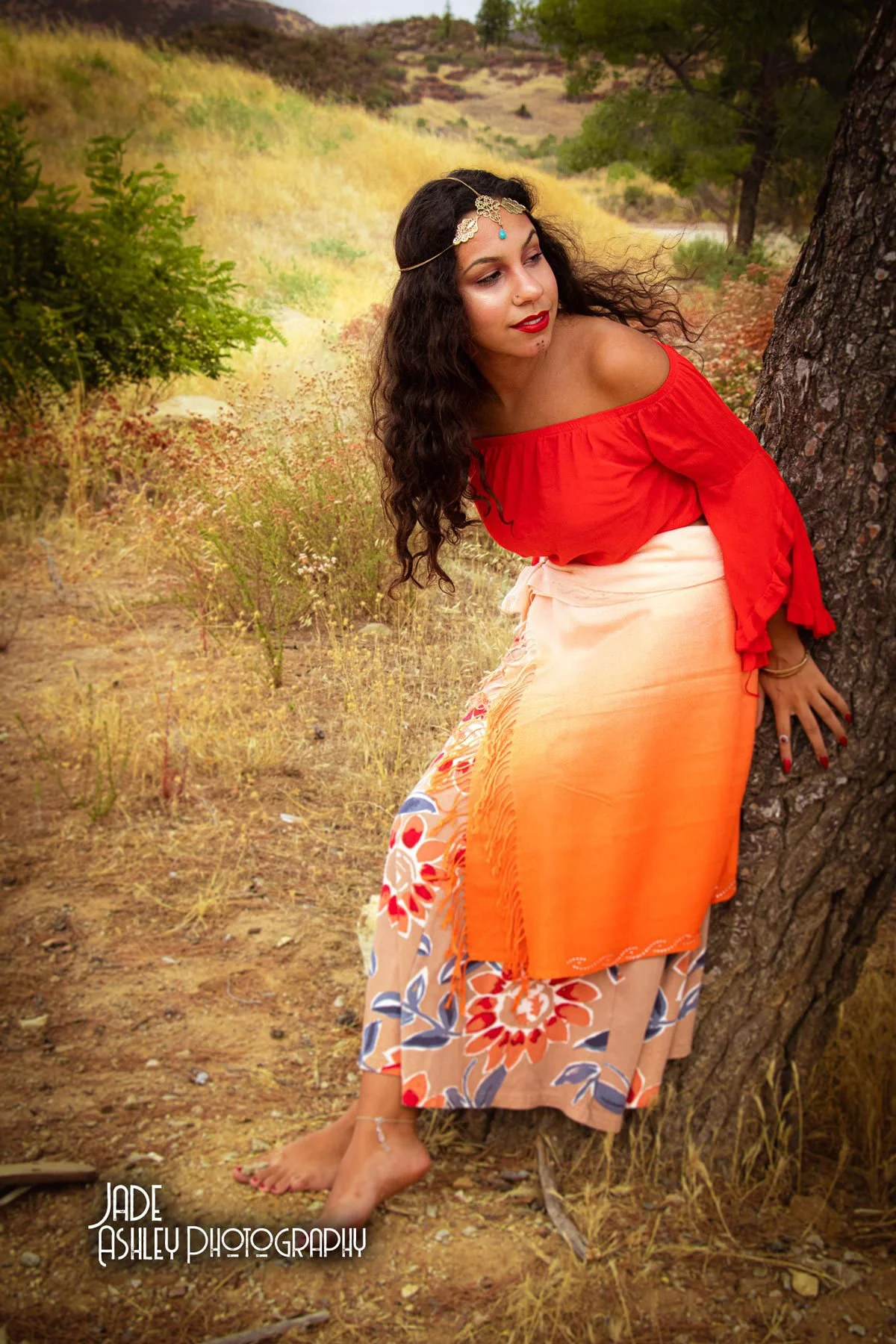 A woman leaning against a tree in a natural outdoor setting, wearing a red off-shoulder top, a long floral patterned skirt, and adorned with jewelry, including a headpiece and bracelet. She has dark, curly hair and is looking to the side with a seren