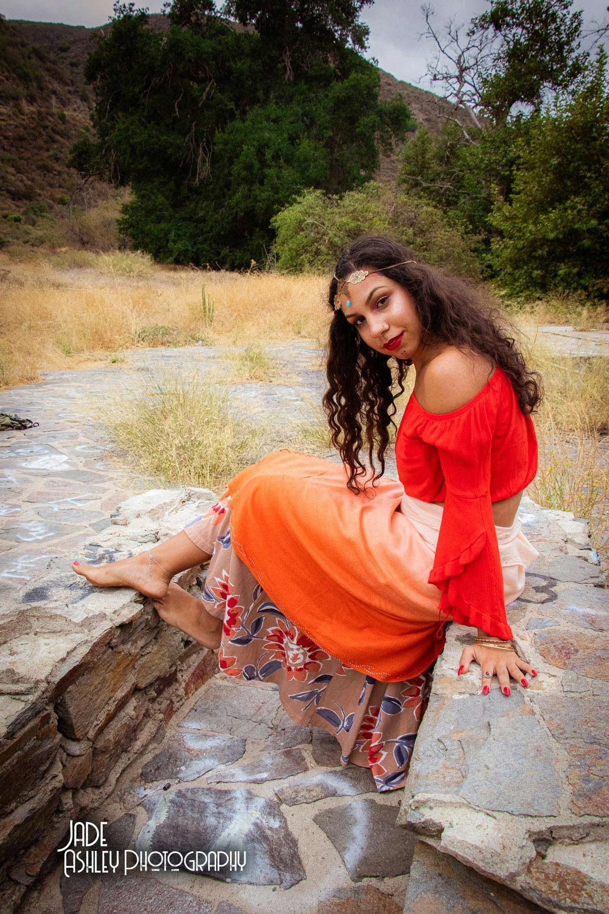A woman in a bohemian style red off-shoulder blouse and a layered skirt with floral patterns, sitting on a stone wall in a dry grassy landscape with trees and hills in the background.