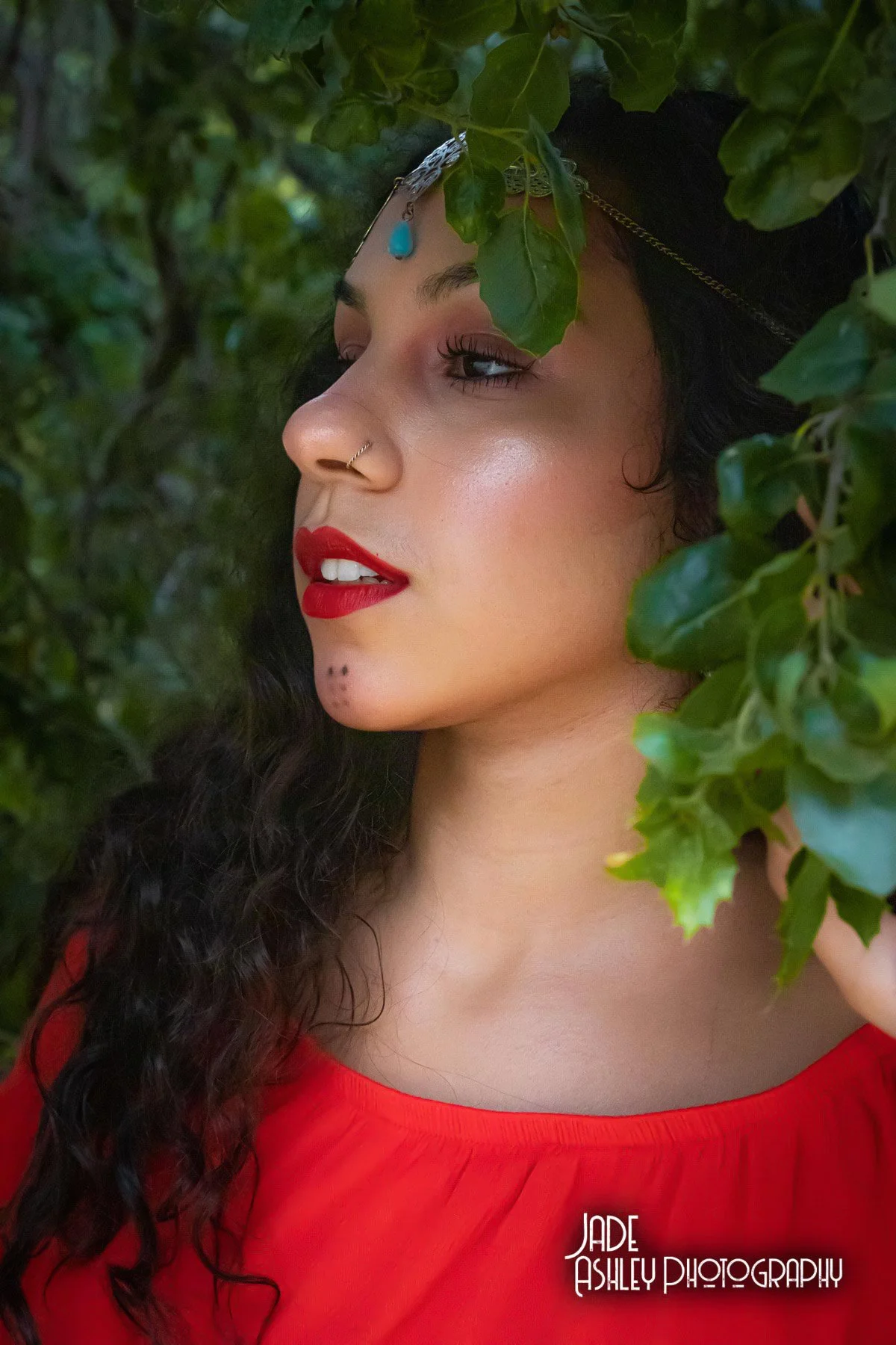 A woman with long curly dark hair and red lipstick, wearing jewelry including a nose ring and headpiece, is surrounded by green leaves, with her face turned to the side.