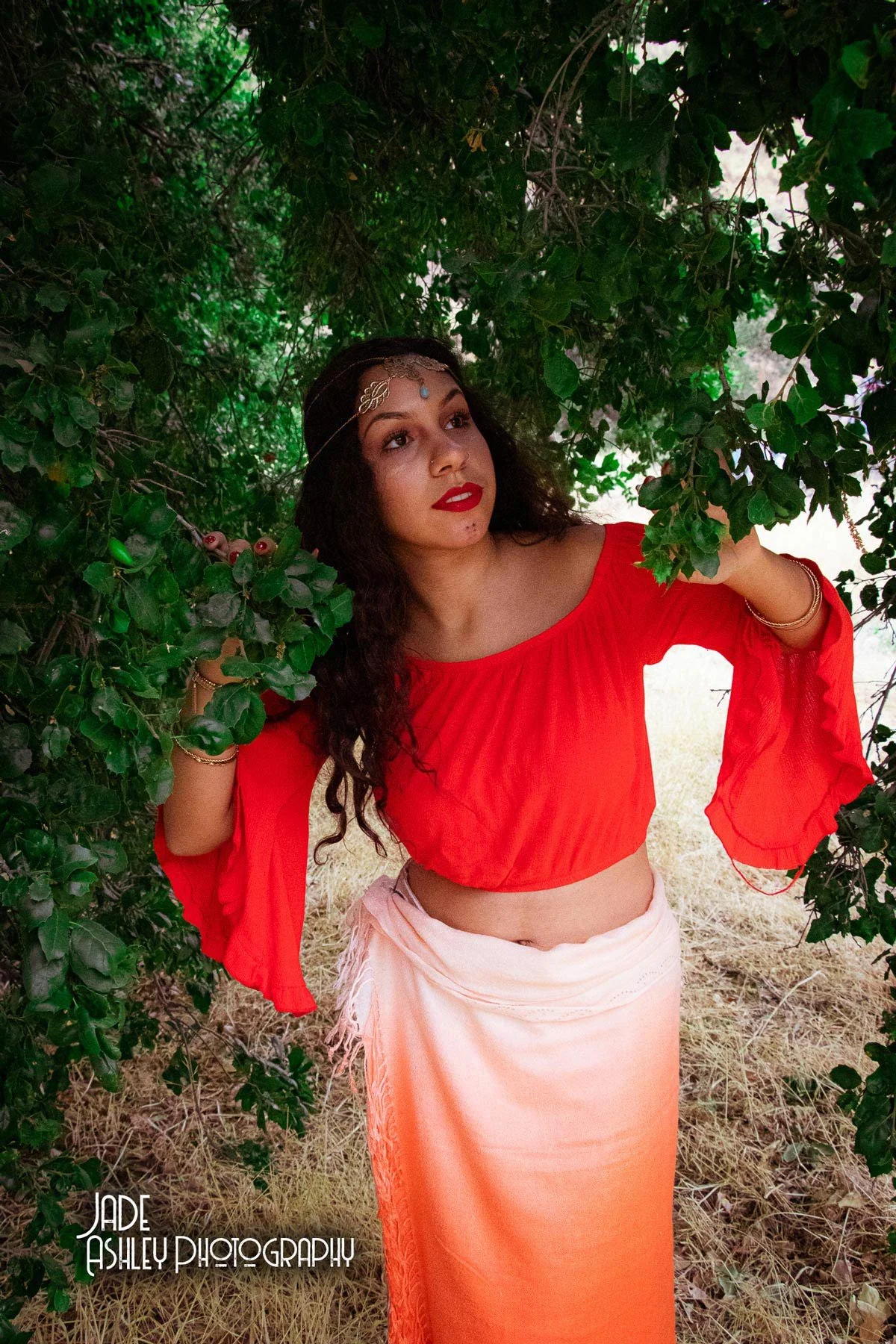 Woman with curly hair and red lipstick wearing a red blouse and peach-colored skirt, standing amid green foliage, looking up and to the side.