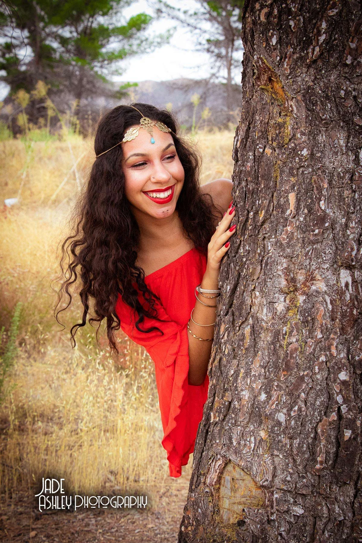 A woman with long, curly dark hair, wearing a red dress and gold jewelry, smiling behind a tree in a field with trees and grass in the background.