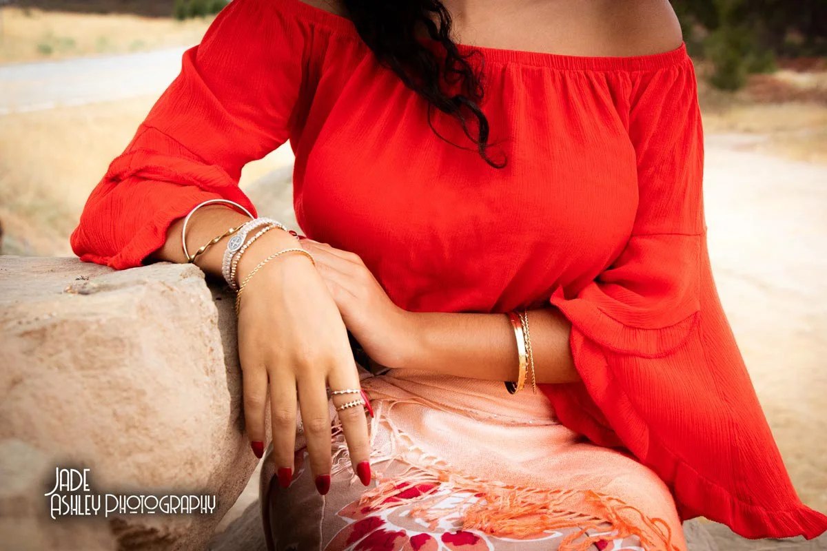 A woman wearing a red off-the-shoulder top and a light patterned skirt, sitting outdoors near a stone surface, with her hands resting on her lap and jewelry adornments on her arms and fingers.