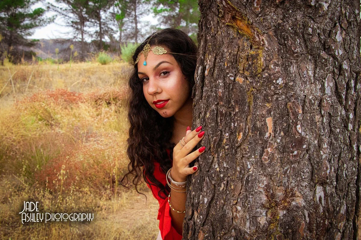 A woman with long, curly dark hair and red lipstick peeks out from behind a tree in a field with dry grass and trees in the background, wearing jewelry and a red outfit.