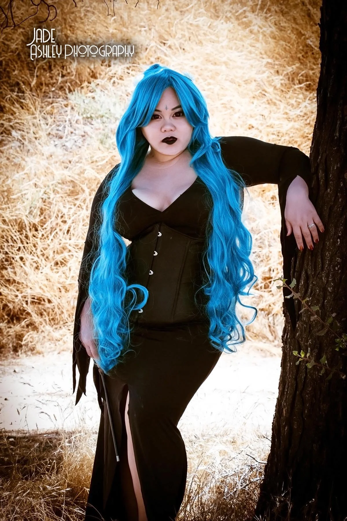 A woman with long, bright blue hair, pale skin, dark lipstick, and face makeup posing outdoors by a tree in a black dress with a slit at the leg, against a background of dry grass and trees.