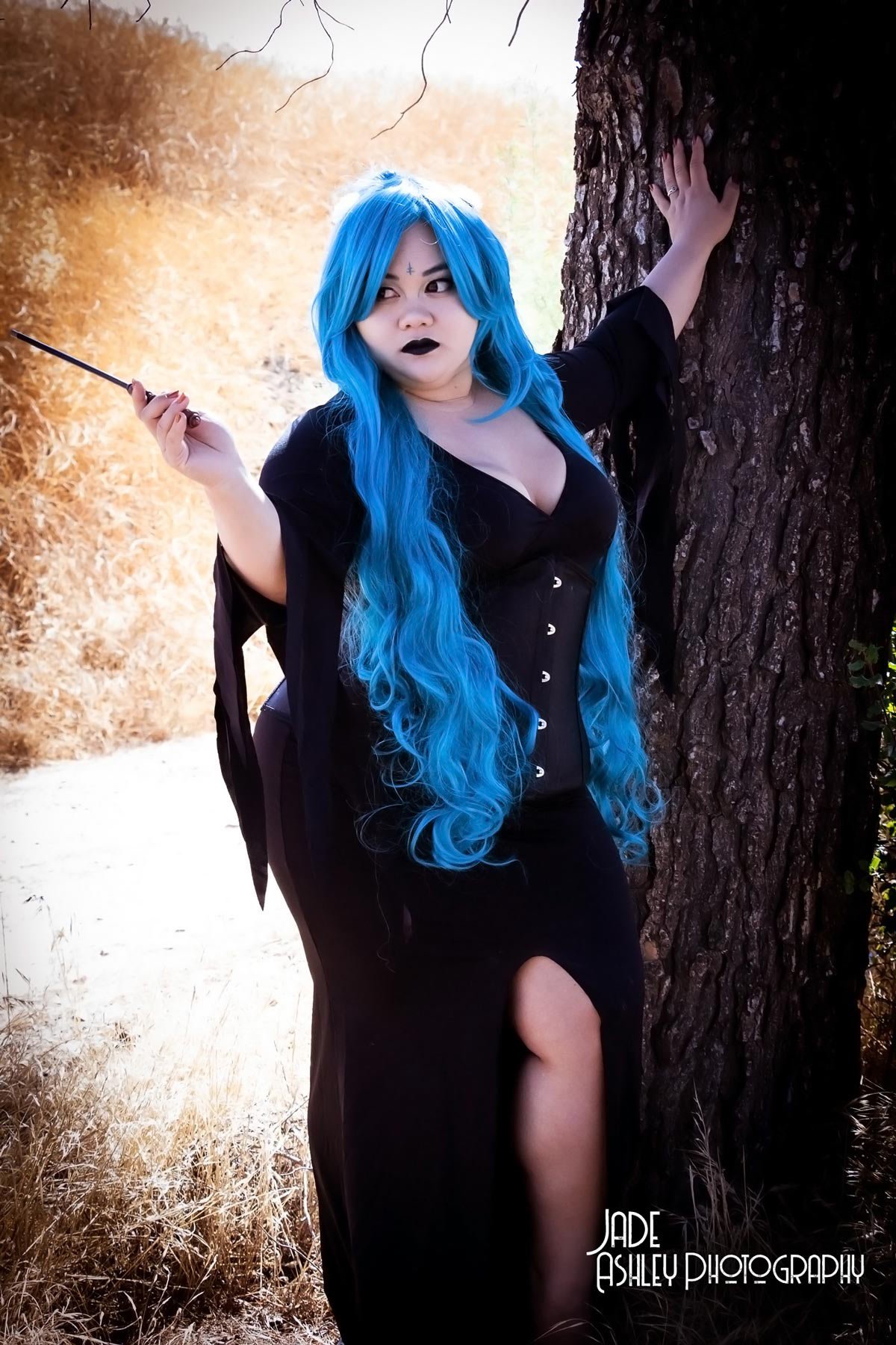 A woman with long blue hair and dark makeup stands outdoors next to a large tree, wearing a black dress with a slit, and holding a small wand or stick in her right hand. The background features a natural landscape with dried grass and trees.
