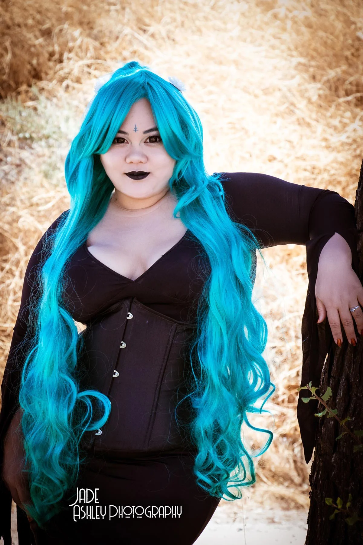 Woman with long, wavy bright blue hair standing outdoors in a natural setting with trees and dry grass, wearing black clothing and dark lipstick.