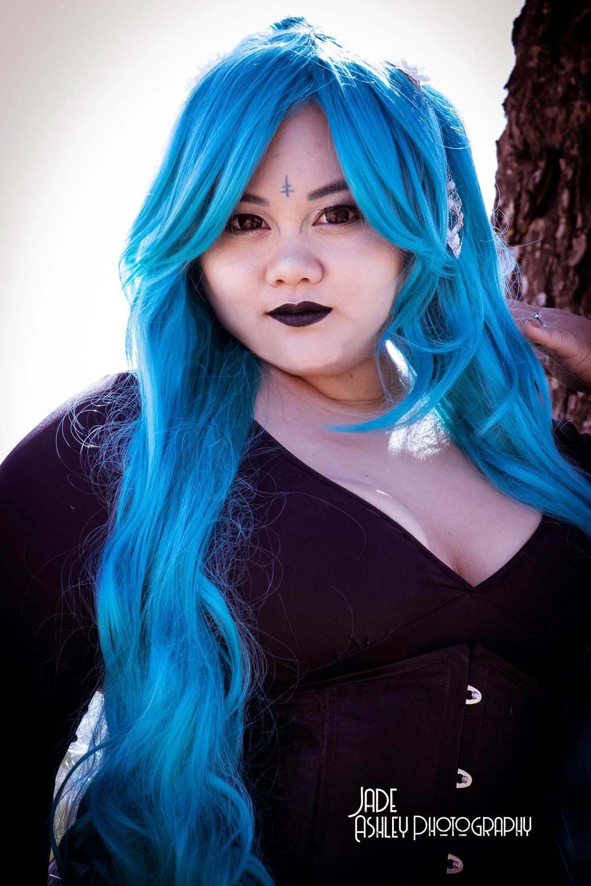A woman with long, wavy, bright blue hair and dark lipstick, standing outdoors next to a tree.