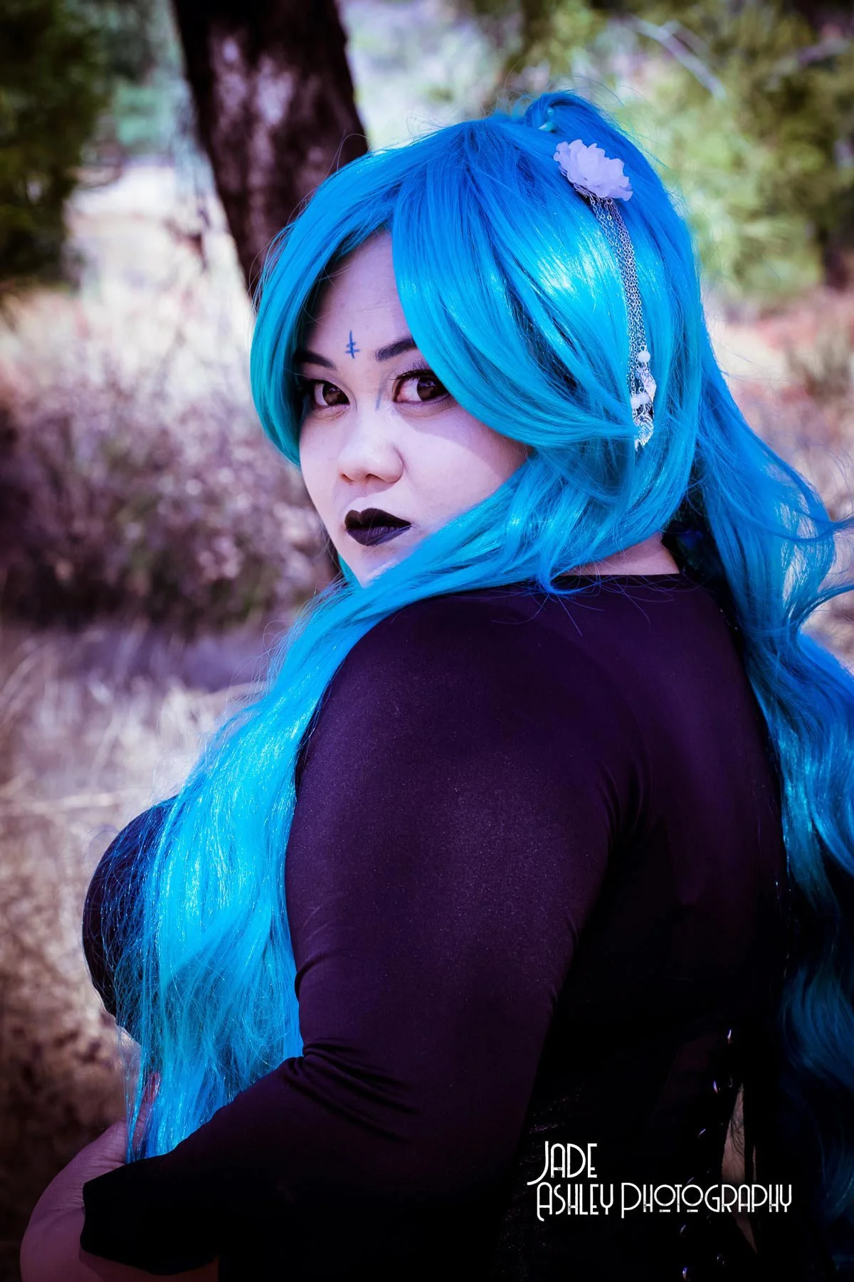 Person with long, bright blue hair, wearing dark makeup, black lipstick, and a jewelry headpiece, standing outdoors with trees and purple-hued foliage in the background.