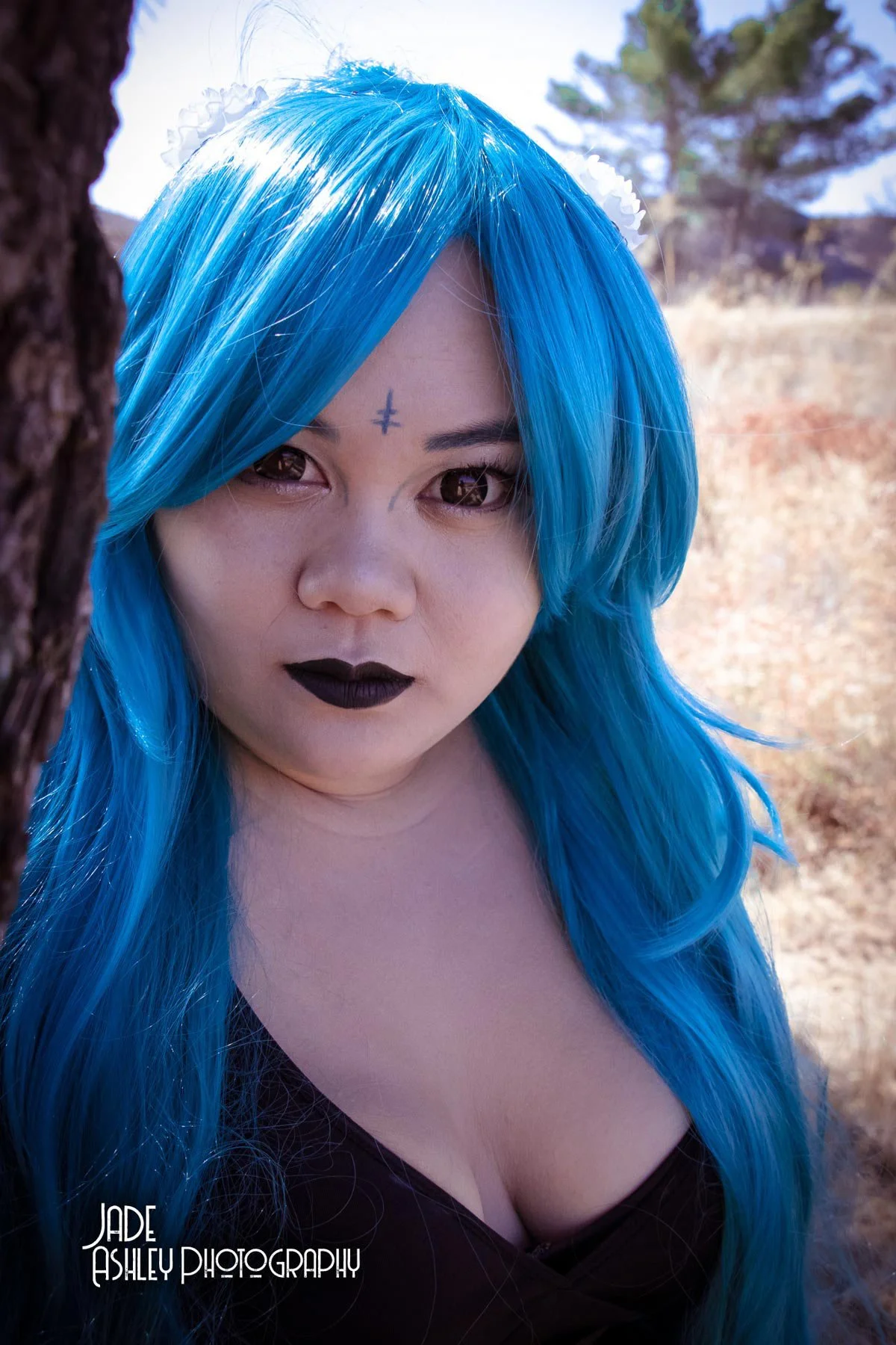 Close-up of a woman with long bright blue hair, dark makeup, and a small tattoo on her forehead, outdoors with trees and dry grass in the background.