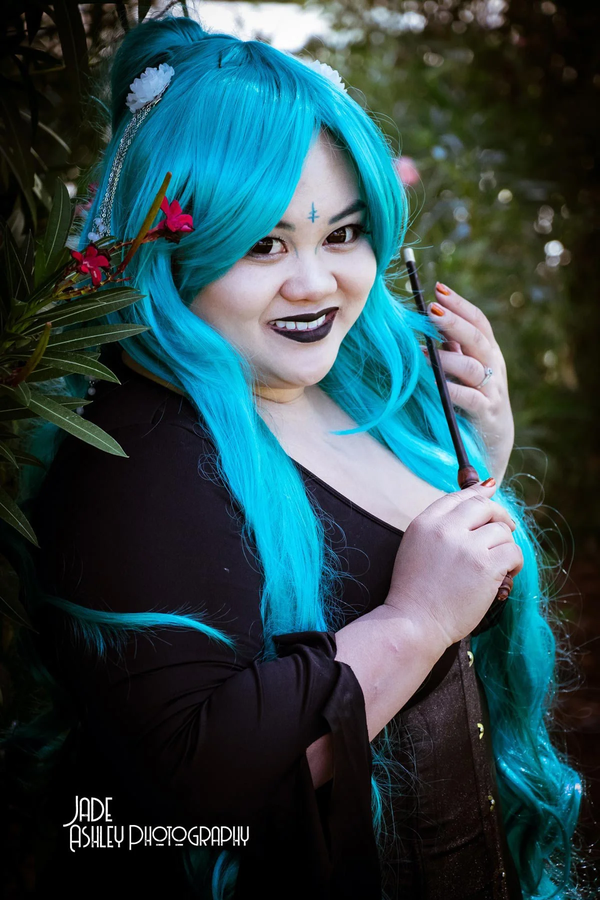 A woman with long, bright turquoise hair holding a black umbrella, smiling while posing outdoors among greenery.