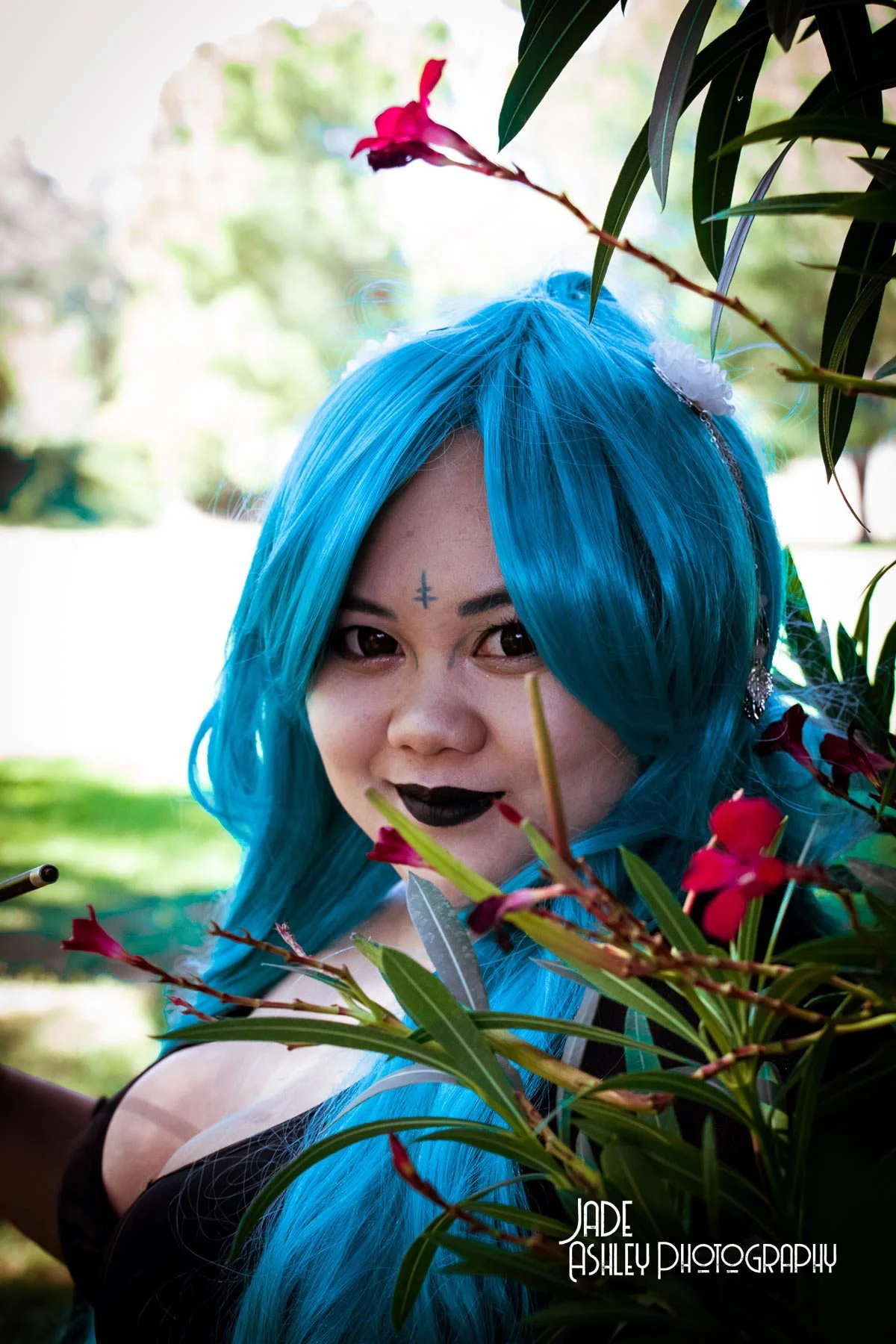 Person with bright blue hair, dark lipstick, and a small tattoo on her forehead peeks through green leaves and red flowers outdoors.