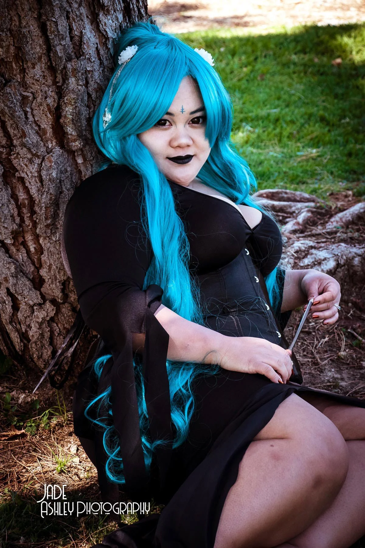 Woman with long blue hair and black lipstick sitting against a tree trunk outdoors, holding a stylus, wearing black clothing and makeup.